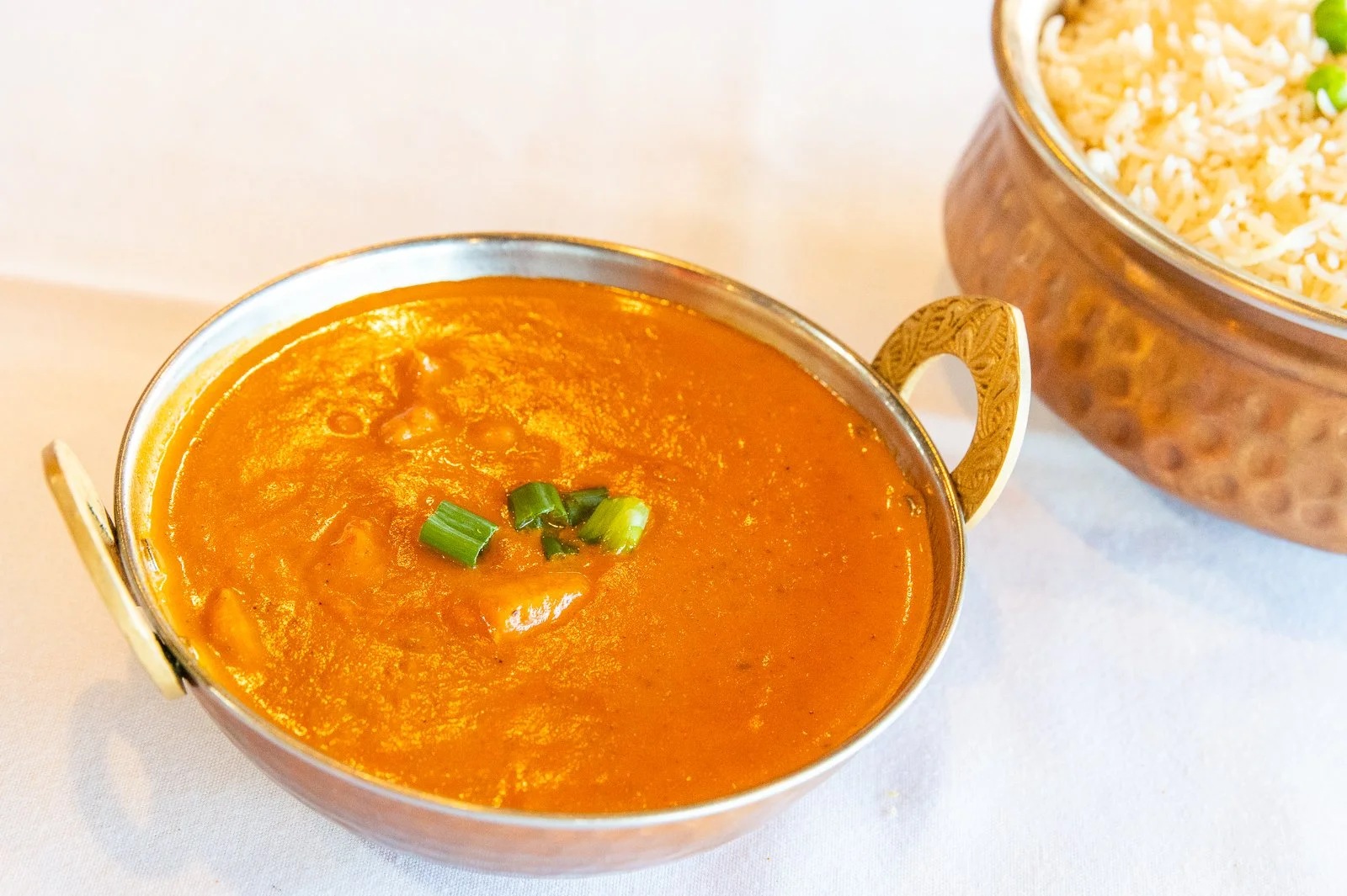 A bowl of orange Indian curry garnished with green onions, next to a bowl of rice