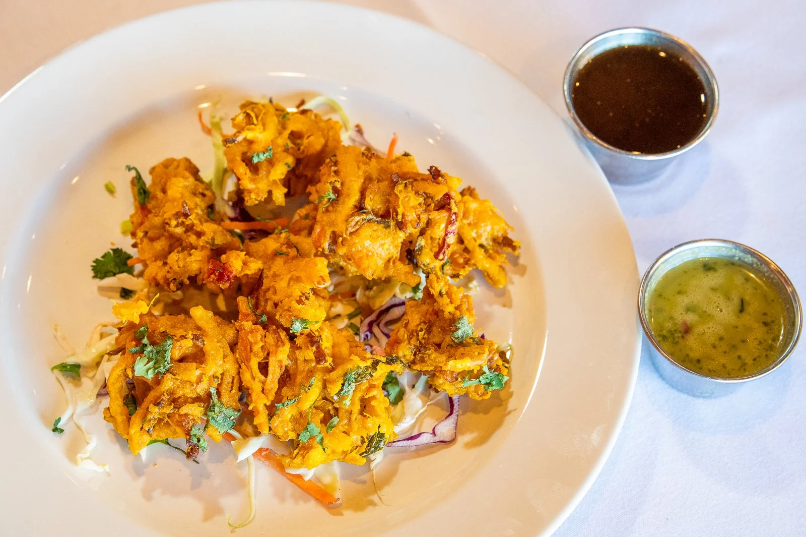 Plate of crispy Indian vegetable fritters garnished with cilantro, accompanied by two small cups of dipping sauces, one dark and one green.