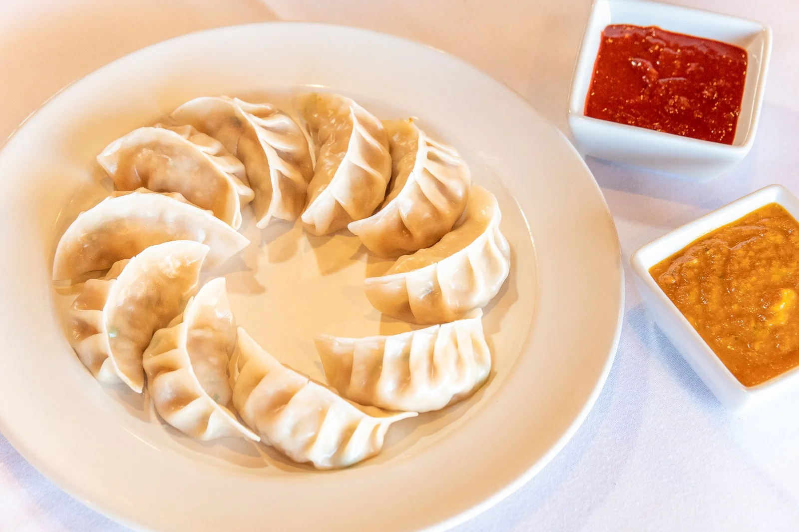 A white plate with Asian dumplings arranged in a circle, two small square bowls of dipping sauces, one red and one yellow, on a white tablecloth.