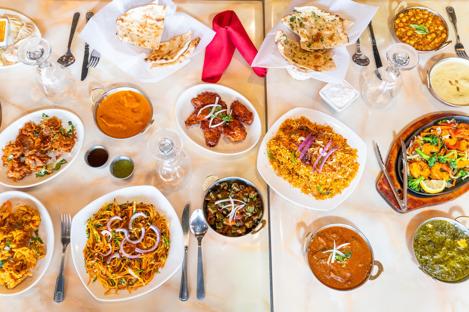 A variety of Indian dishes on a table including naan bread, chicken wings, fried rice, noodles, vegetable curry, lentil soup, and various sauces, served on white plates and bowls with glasses and cutlery.