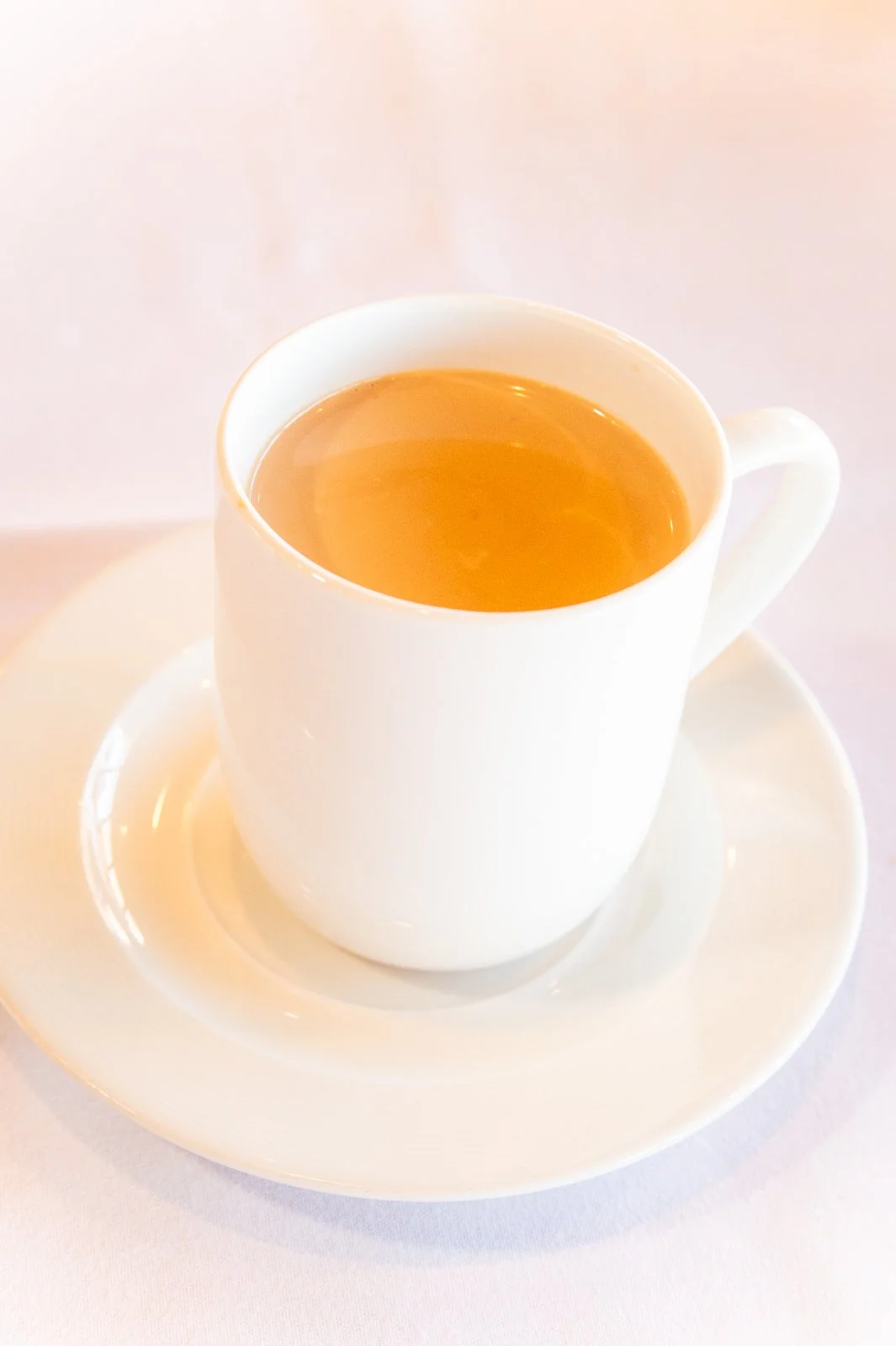A white ceramic cup filled with tea on a matching saucer, placed on a light-colored table.