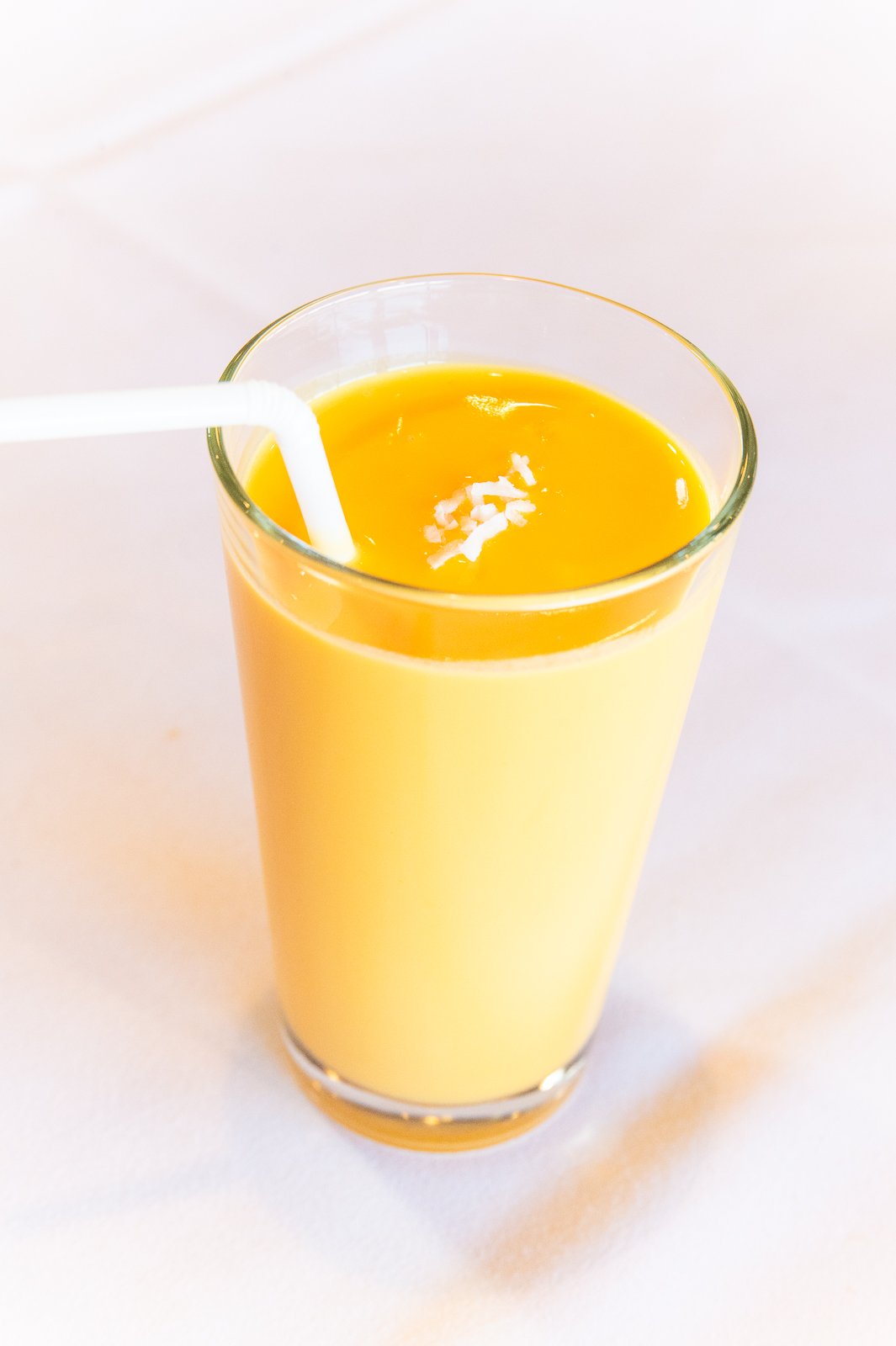 A tall glass filled with yellow mango juice, garnished with small white bits of coconut or pineapple, with a white straw, on a white surface.