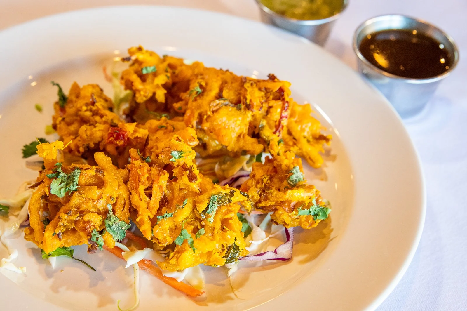 Fried Indian vegetable fritters served on a white plate with cabbage and carrots, garnished with cilantro, accompanied by small bowls of green chutney and tamarind sauce.