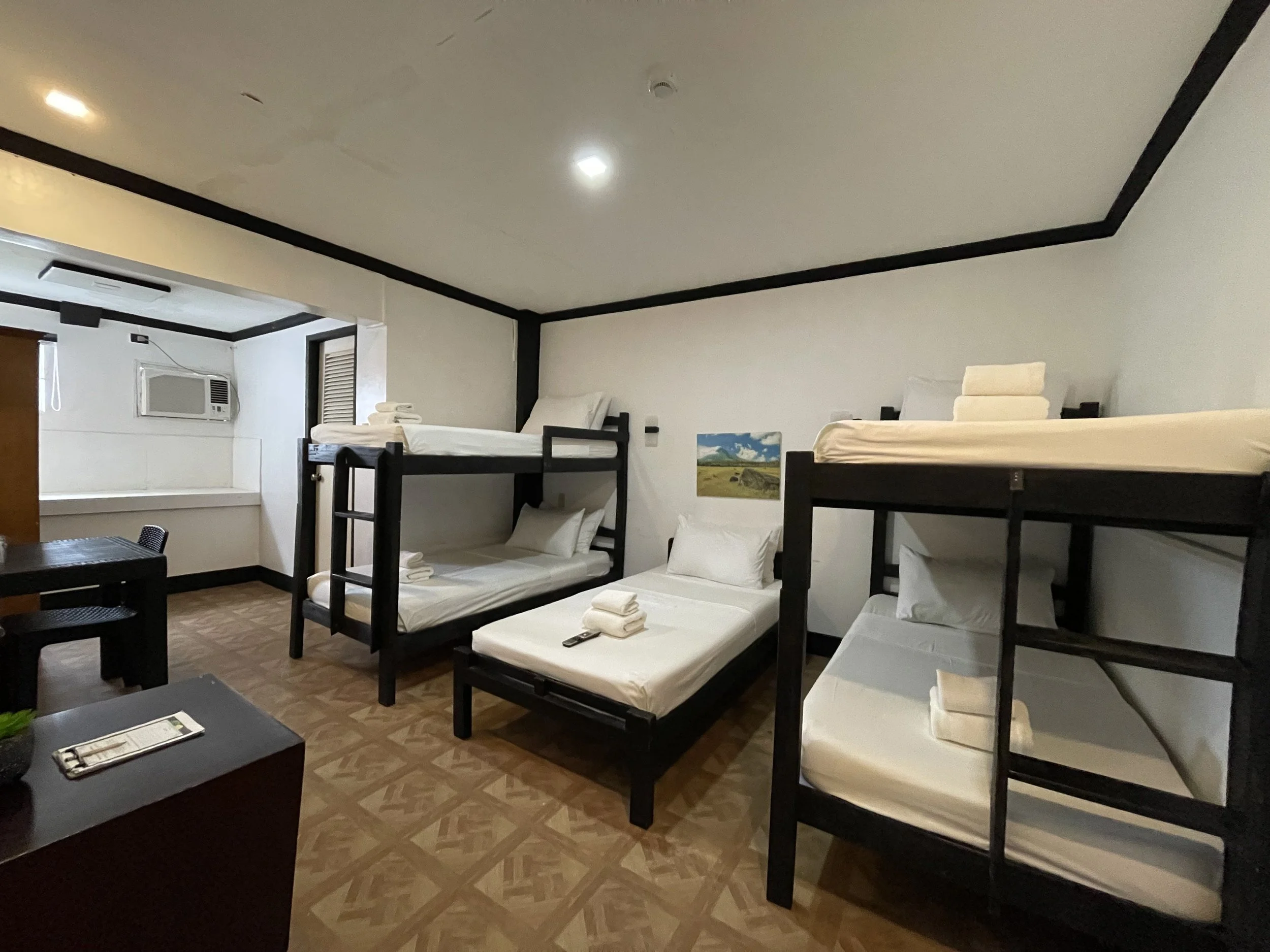 Room with two sets of bunk beds, each with two beds, towels, pillows, and a small painting on the wall, with a window air conditioning unit and a table with chairs.