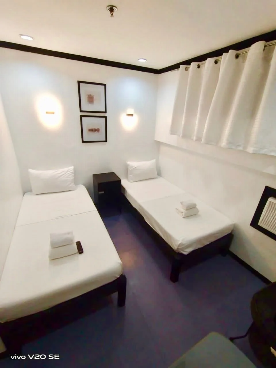 A small hotel room with two single beds, white bedding, and pillows. There is a black nightstand between the beds, framed pictures on the wall, two wall-mounted lights, and a window with cream-colored curtains.