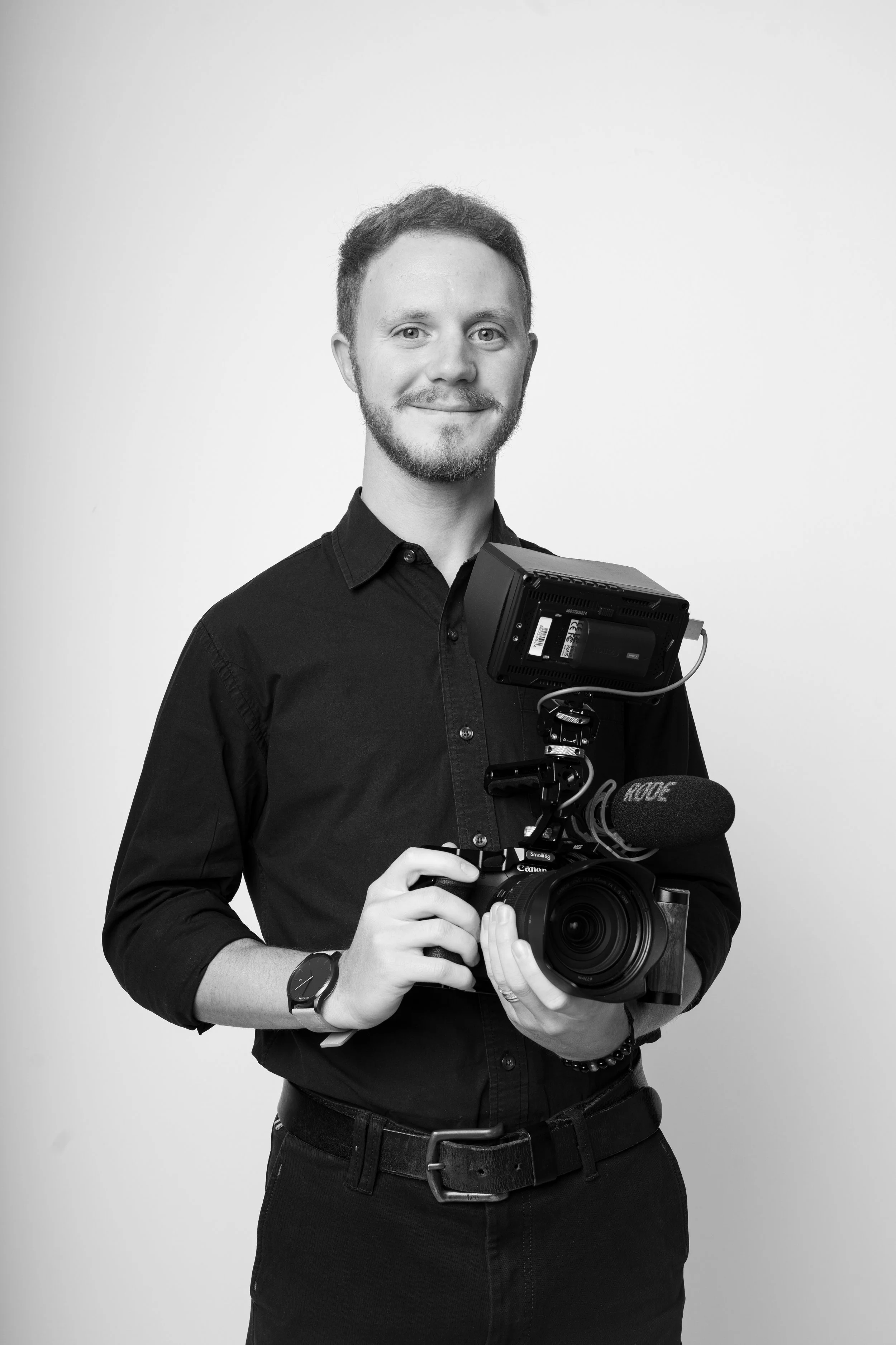 Uomo che tiene una videocamera professionale con microfono e schermo, indossa un orologio e si trova in uno studio con sfondo bianco.