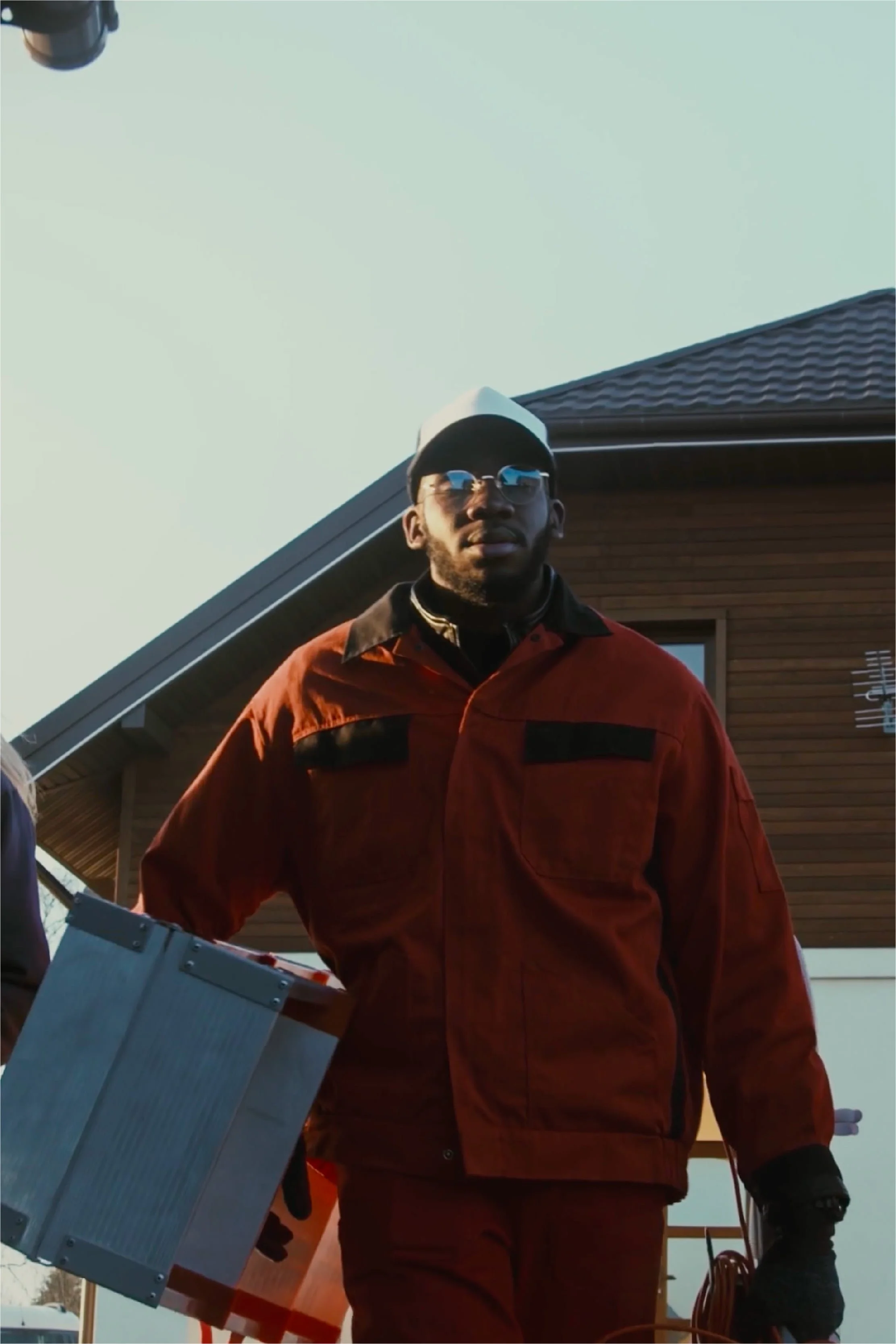 Homme portant une veste rouge, des lunettes de soleil, un bonnet blanc et noir, tenant une boîte métallique, debout devant une maison en bois et un ciel dégagé.
