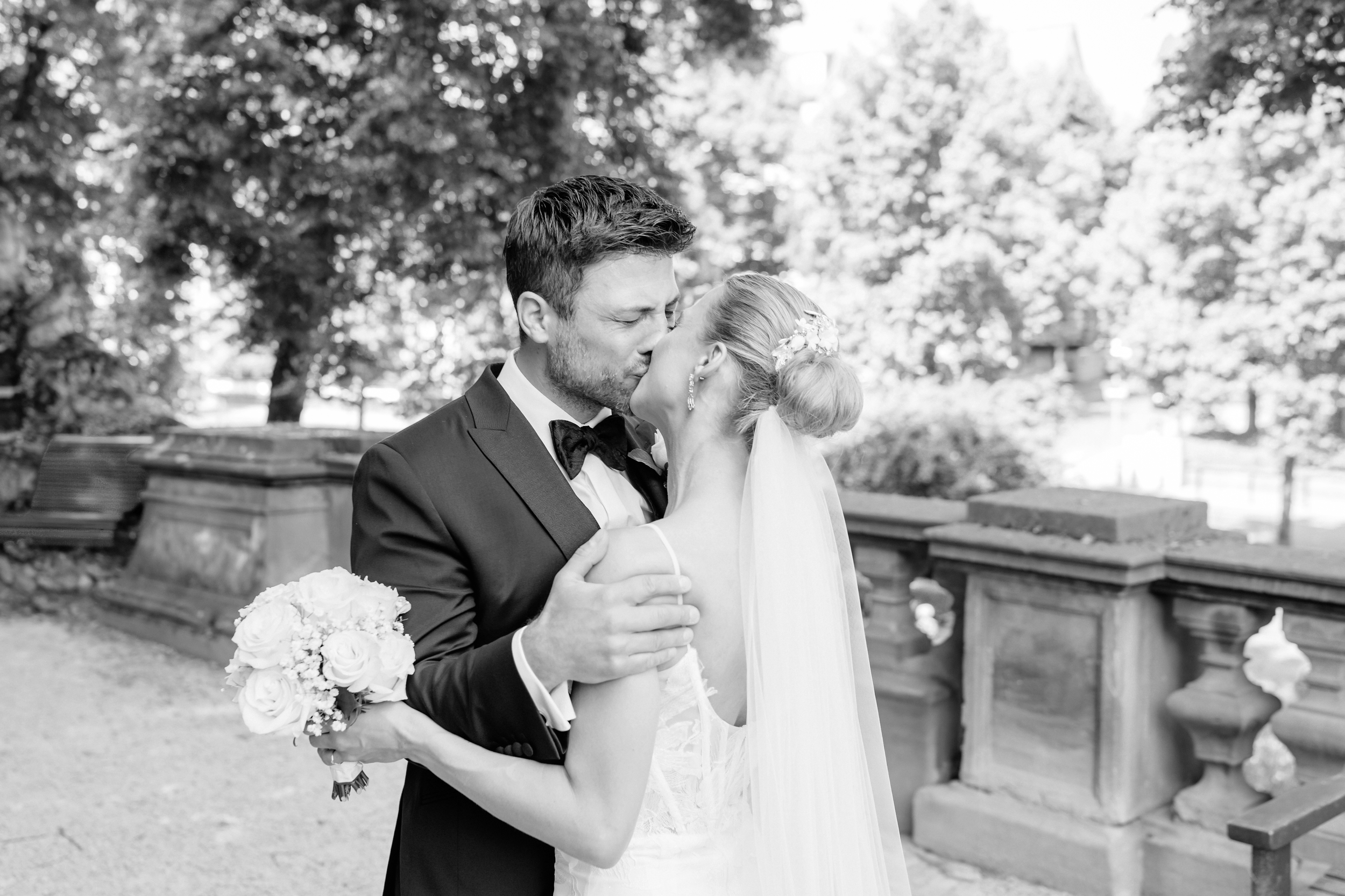 Ein Brautpaar teilt einen Kuss im Freien, die Braut hält einen Blumenstrauß, im Hintergrund Bäume und eine steinerne Balustrade. Sven Goos Fotografie