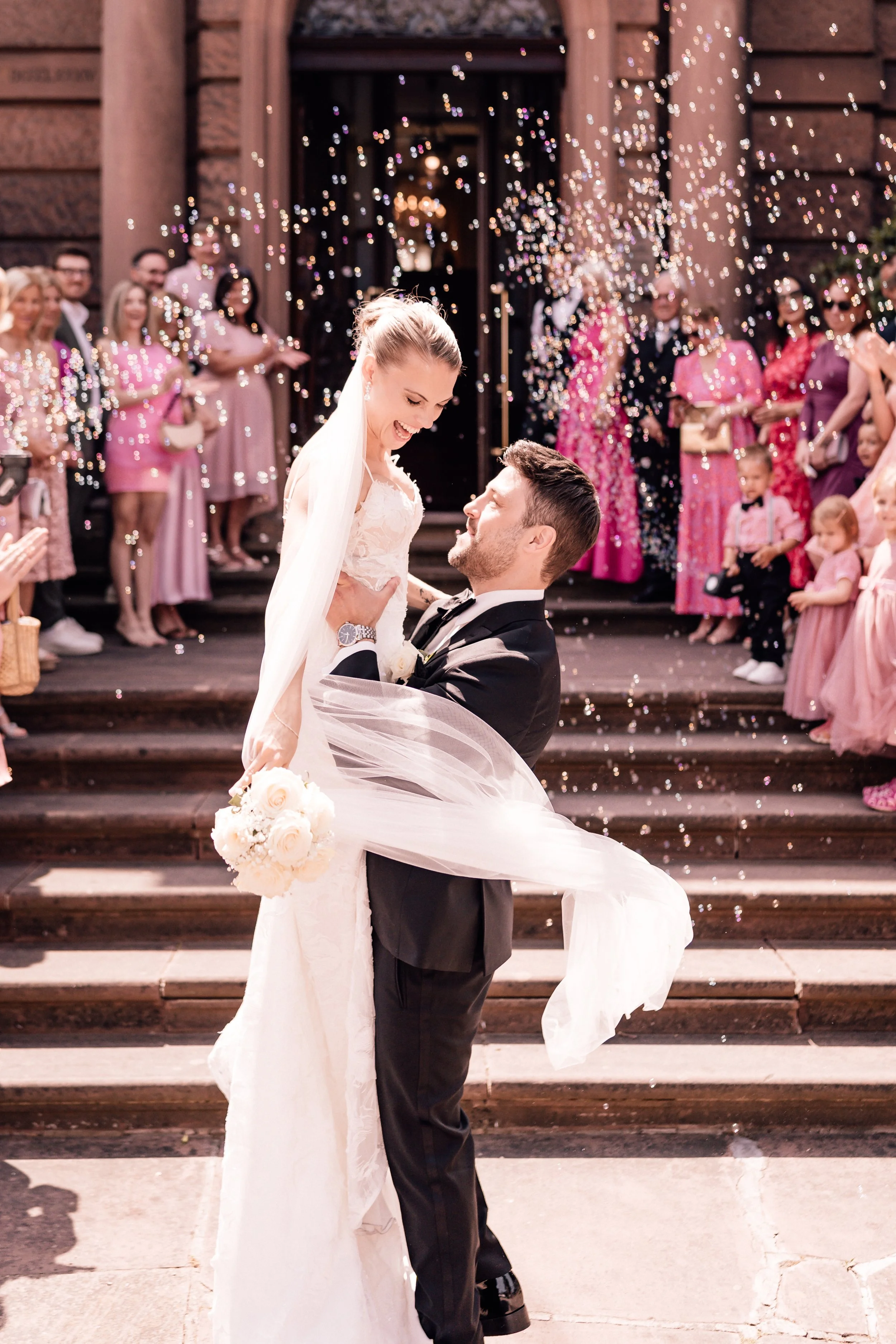 Ein glücklich verheiratetes Paar, das sich auf den Stufen einer Kirche freut, umgeben von Gästen, die mit Konfetti werfen, bei einer Hochzeitsfeier. Sven Goos Fotografie