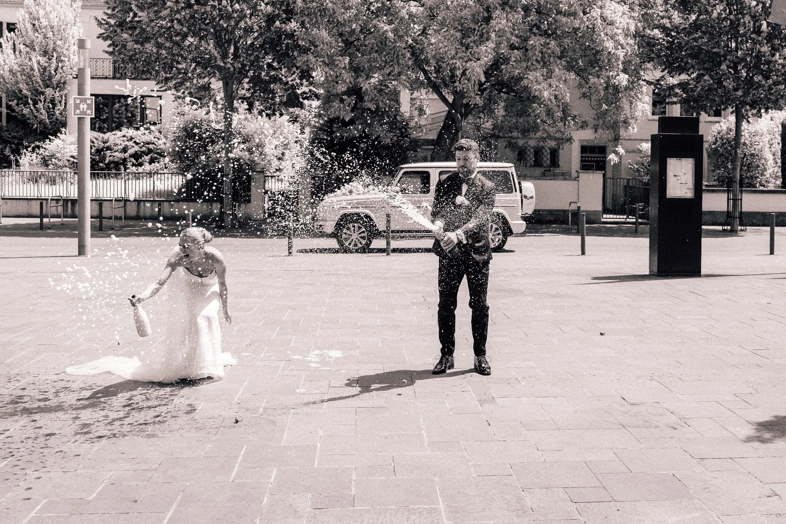 Ein Mann und ein junges Mädchen, die im Freien auf einer gepflasterten Fläche mit einer Wasserpistole Wasser spritzen. Im Hintergrund sind Bäume, ein Auto und eine Werbetafel sichtbar. Sven Goos Fotografie