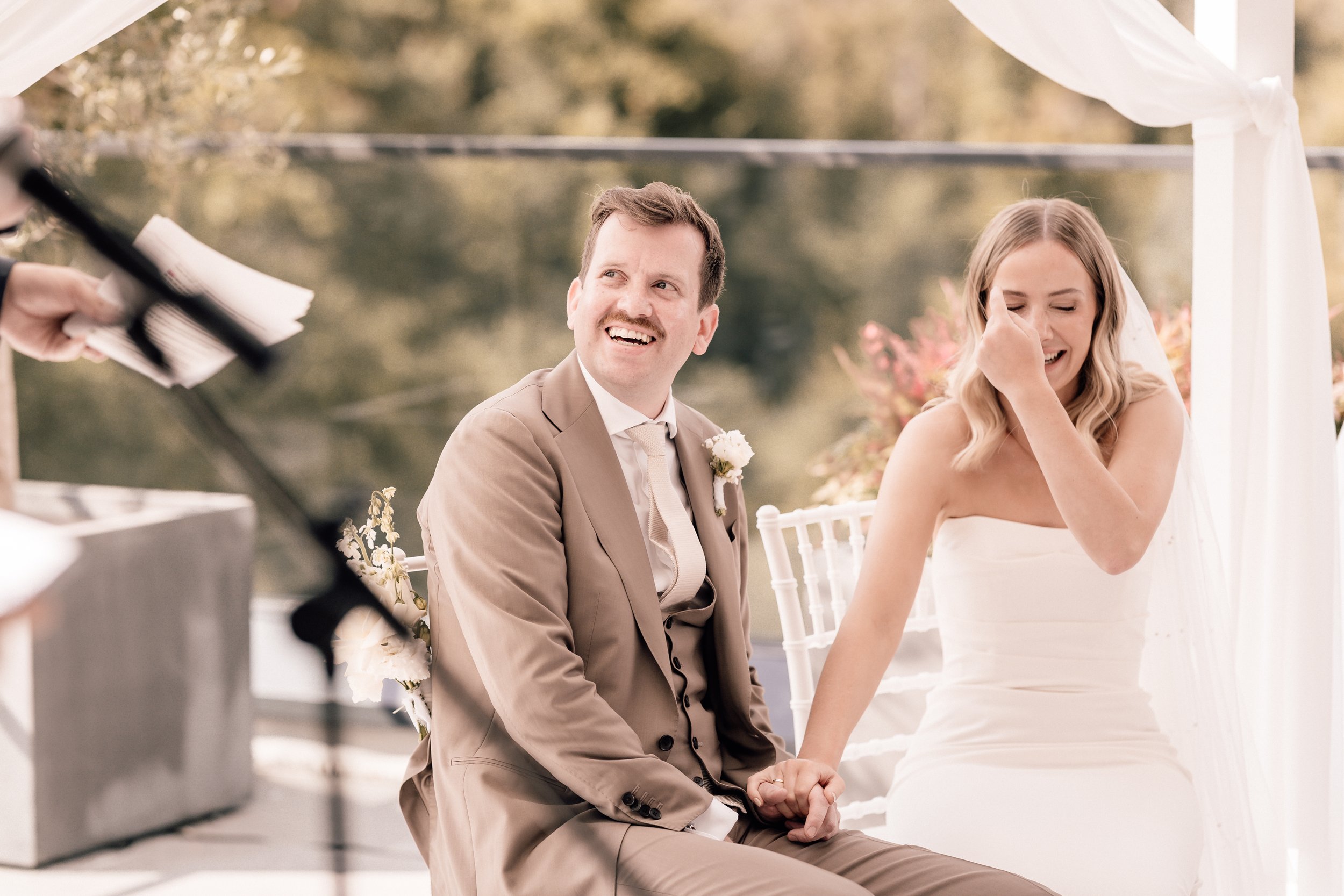 Ein Brautpaar bei der Trauung im Freien, der Bräutigam lacht, die Braut ist emotional berührt und weint, während sie an einem Tisch sitzen, umgeben von Natur und Blumen. Sven Goos Fotografie