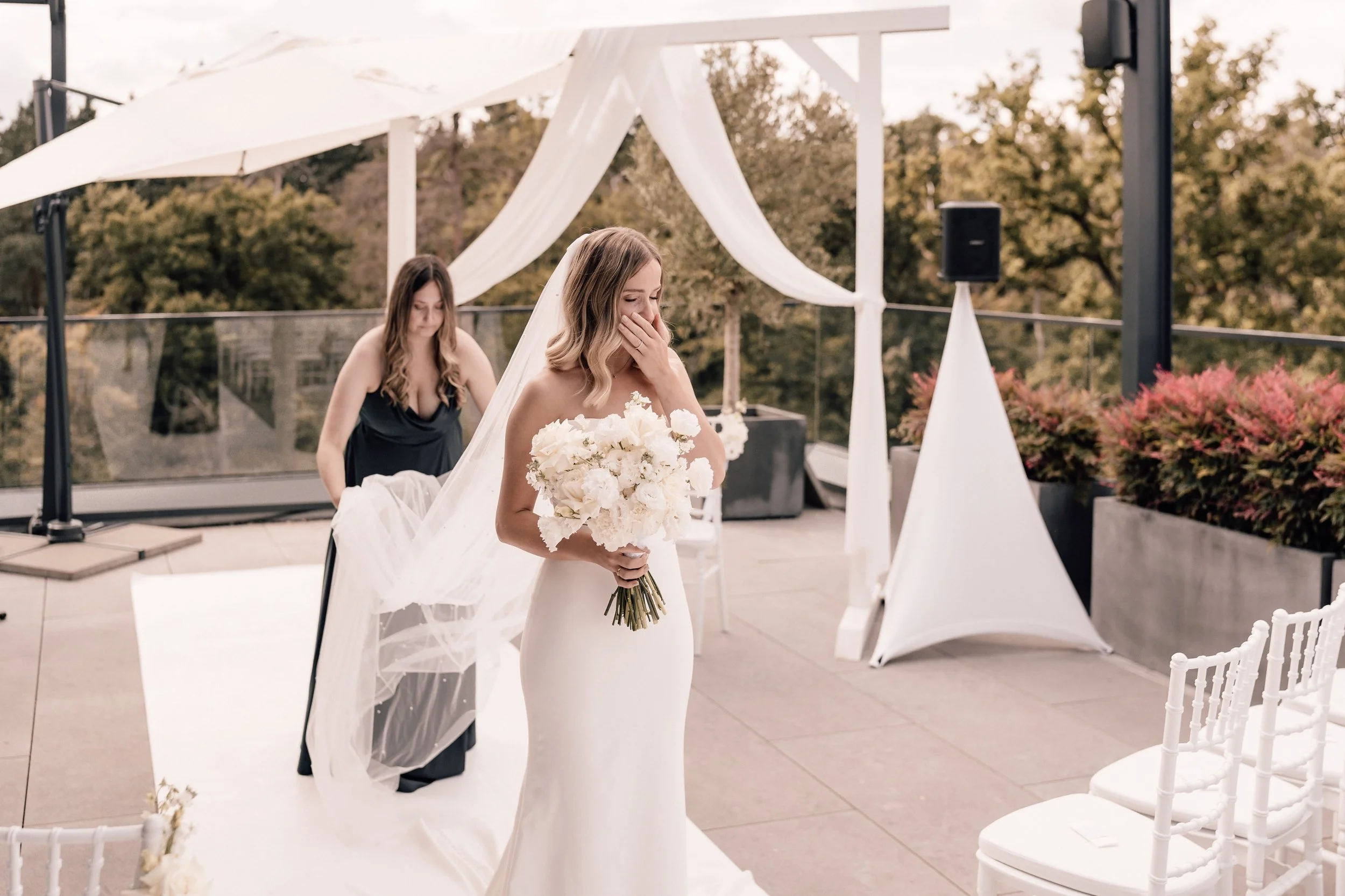 Braut in weißem Hochzeitskleid mit Blumenstrauß, die in einer Dekoration auf einem Dachgarten steht, während eine Helferin im Hintergrund die Brautkleid-Anprobe vorbereitet und Gurten befestigt. Sven Goos Fotografie