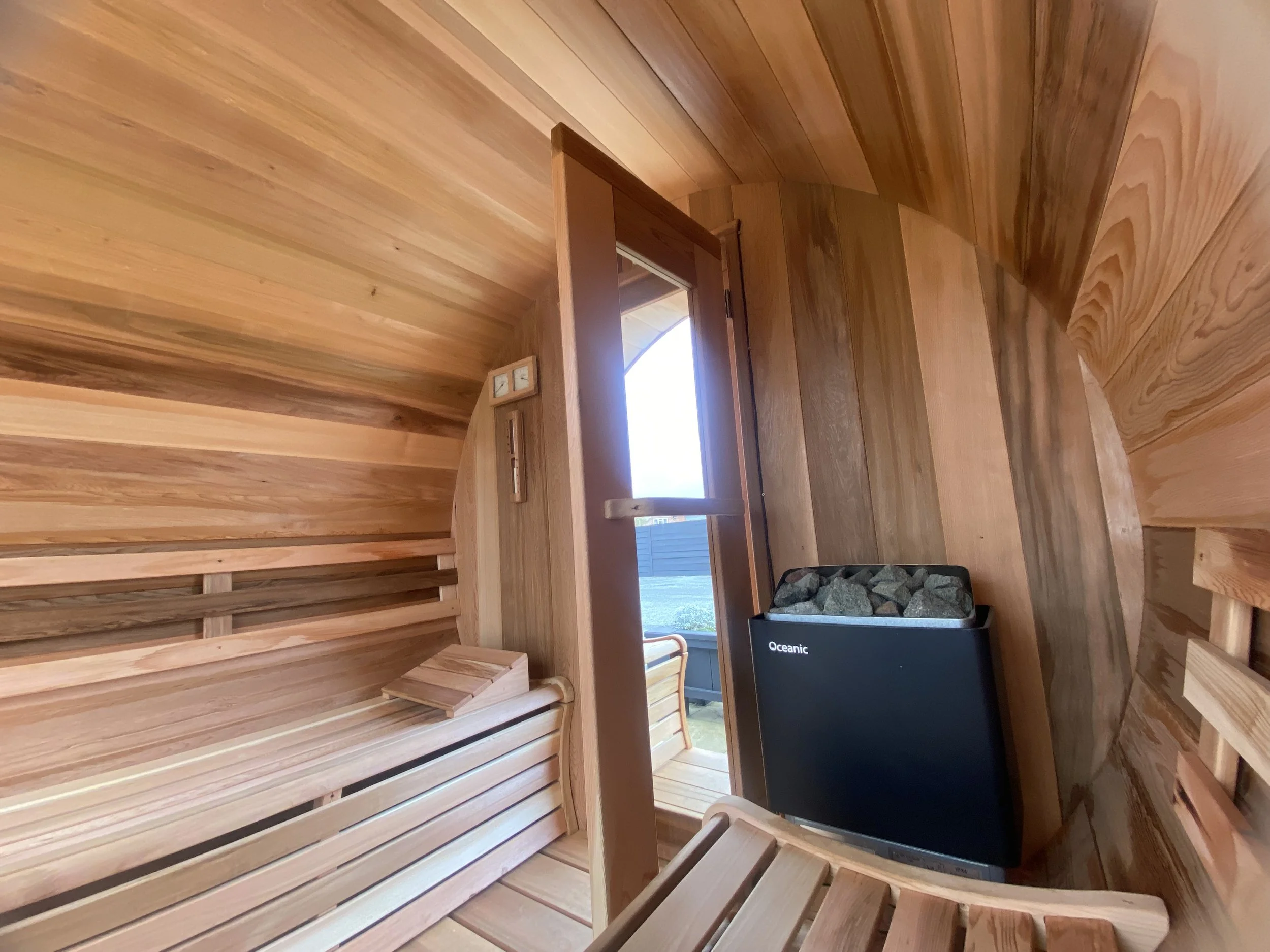 Interior of a small wooden sauna with benches, a heater with rocks, and a view outside through an open door.