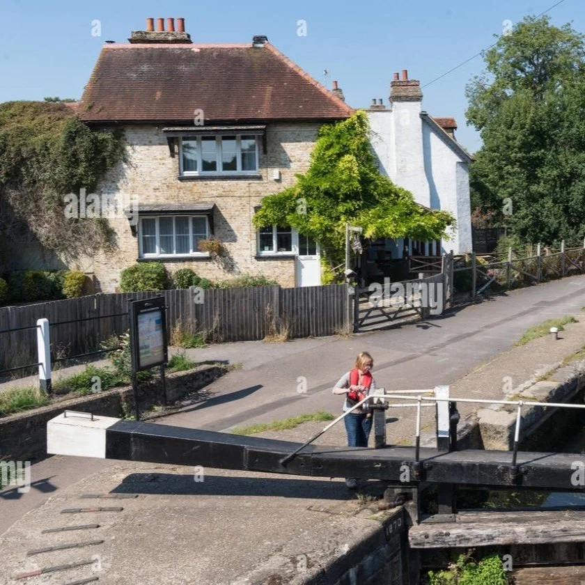 A woman with a red backpack leaning on a lock gate of a canal near a charming house with a sloped roof and brick walls, surrounded by greenery.