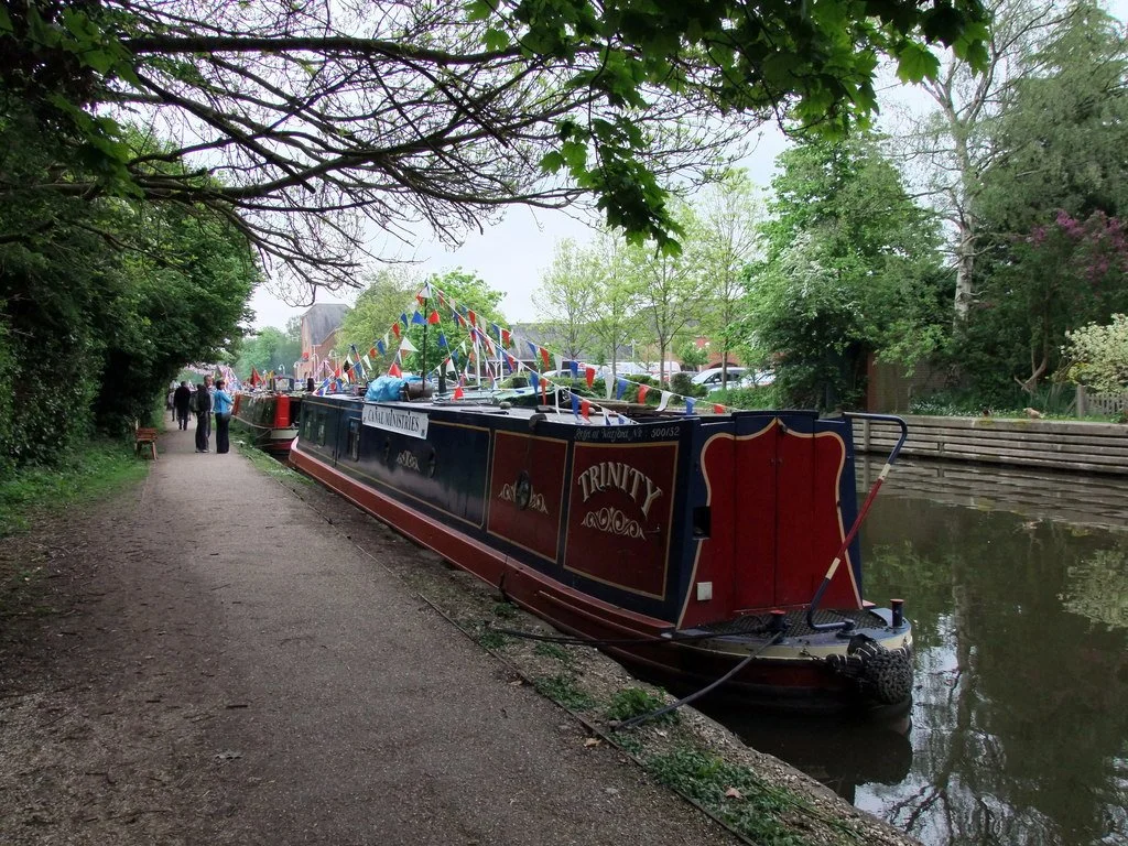 A narrowboat decorated for a festival, floating on a canal next to a walk path with trees, flowering bushes, and people in the background.