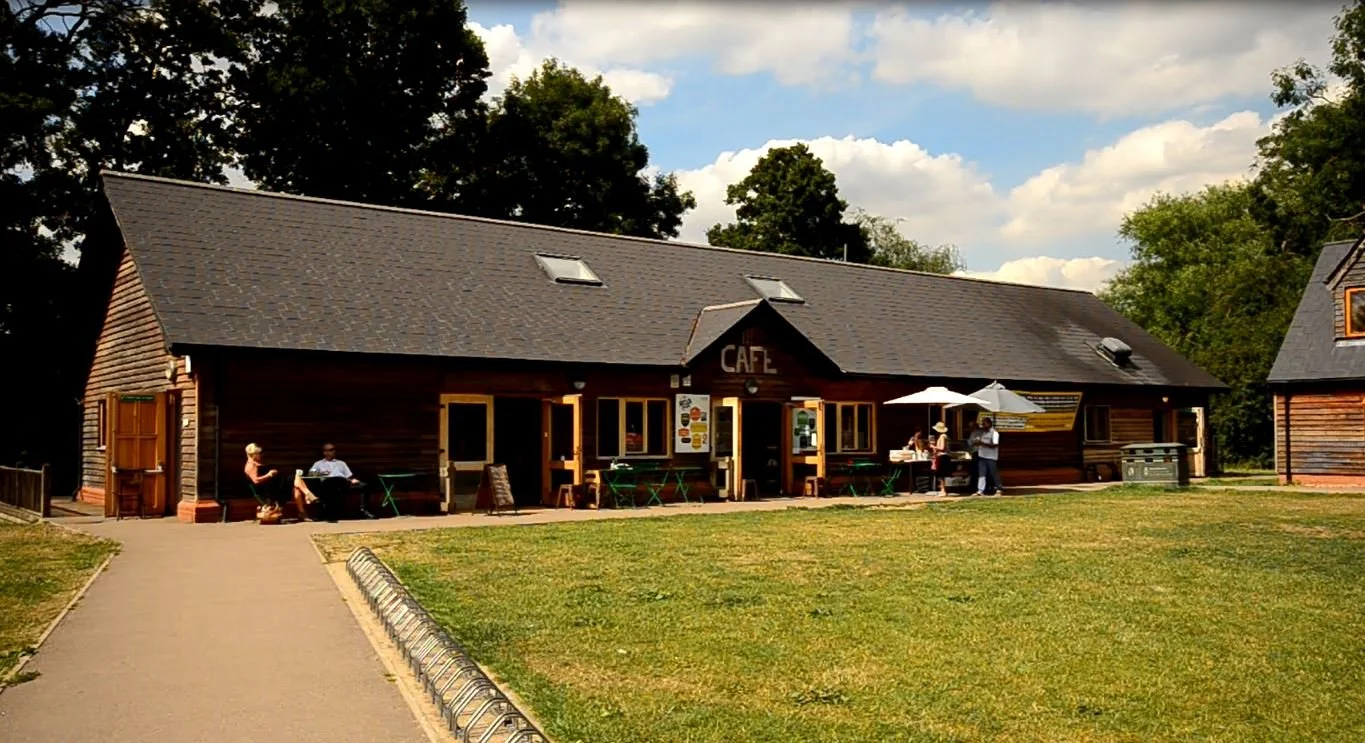 Wooden cafe building with outdoor seating, people sitting under umbrellas, green lawn, and trees in the background.