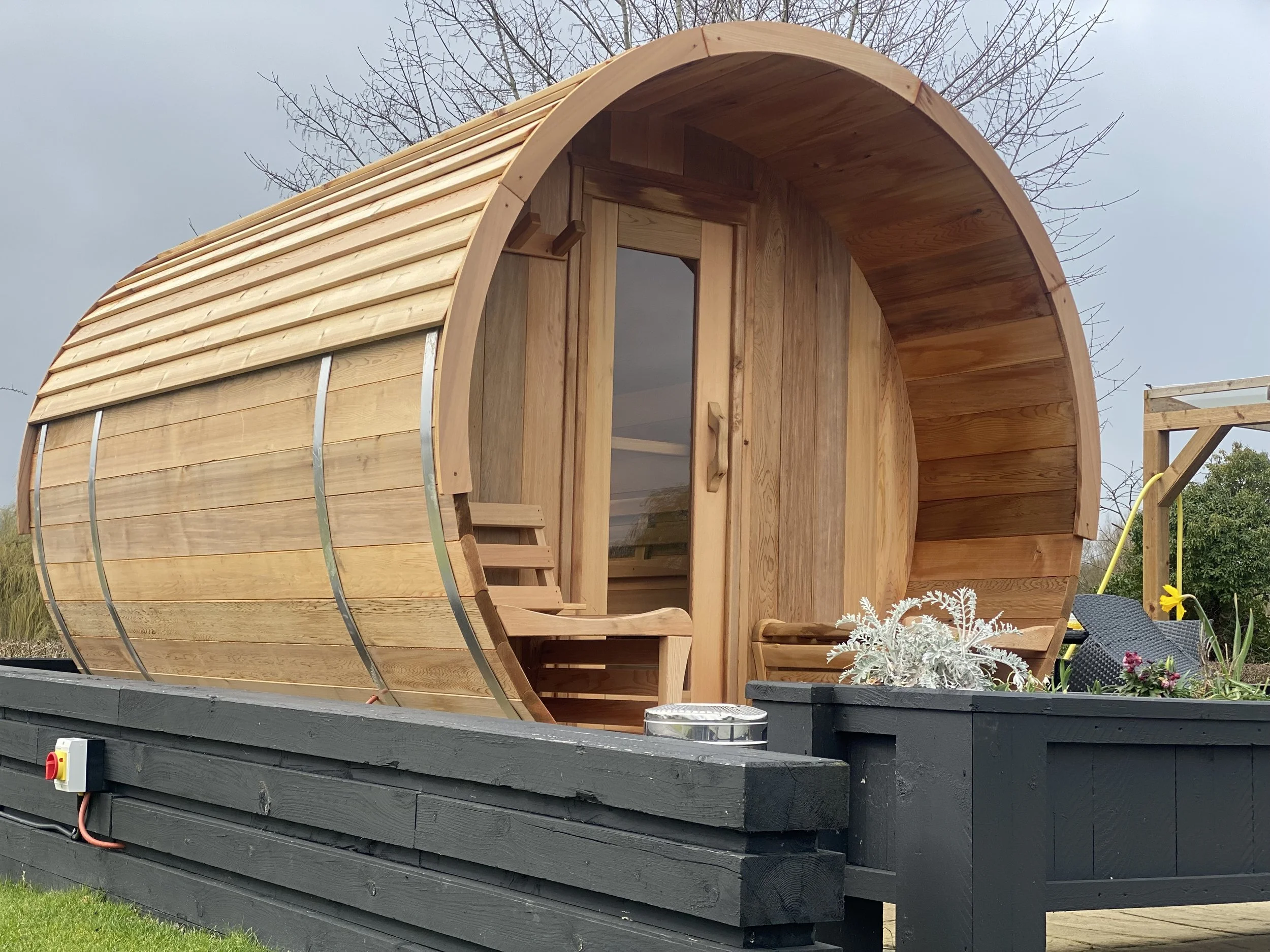 A wooden barrel-shaped sauna on a black platform with plants and outdoor scenery in the background.