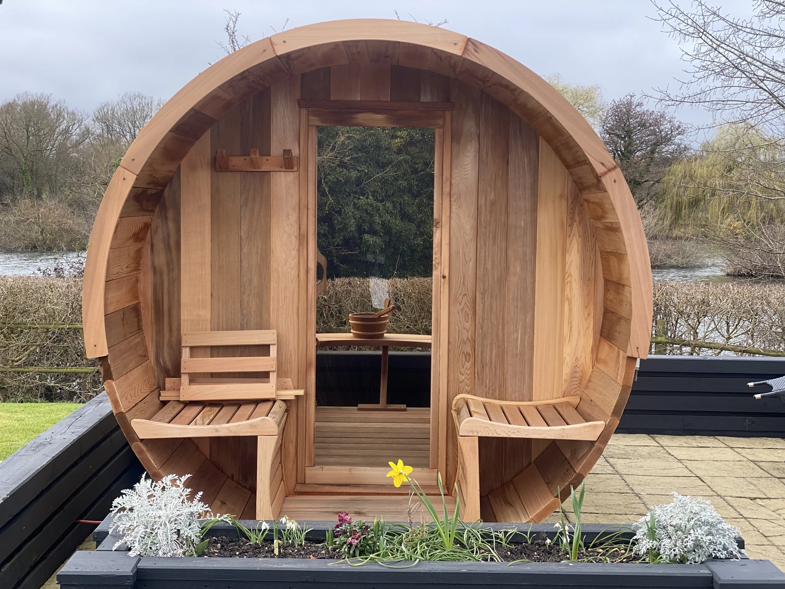 Round wooden hot tub with benches inside, outside on patio with plants and river in the background.