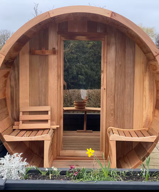 A round wooden sauna with a door opening to an outdoor view of trees, featuring wooden benches inside and some flowering plants outside.