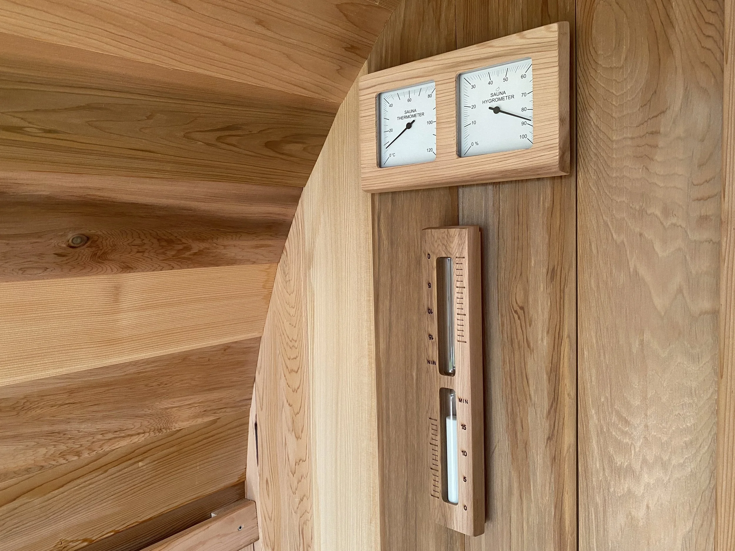 A wooden sauna wall with two gauges, a thermometer and a hygrometer, along with a wooden hydrometer, measuring temperature and humidity levels.