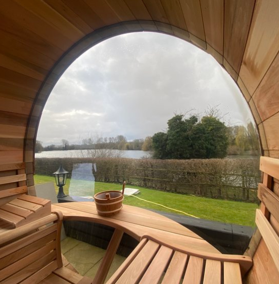 View through a large, round window on a wooden sauna showing a lake, trees, and cloudy sky outside.