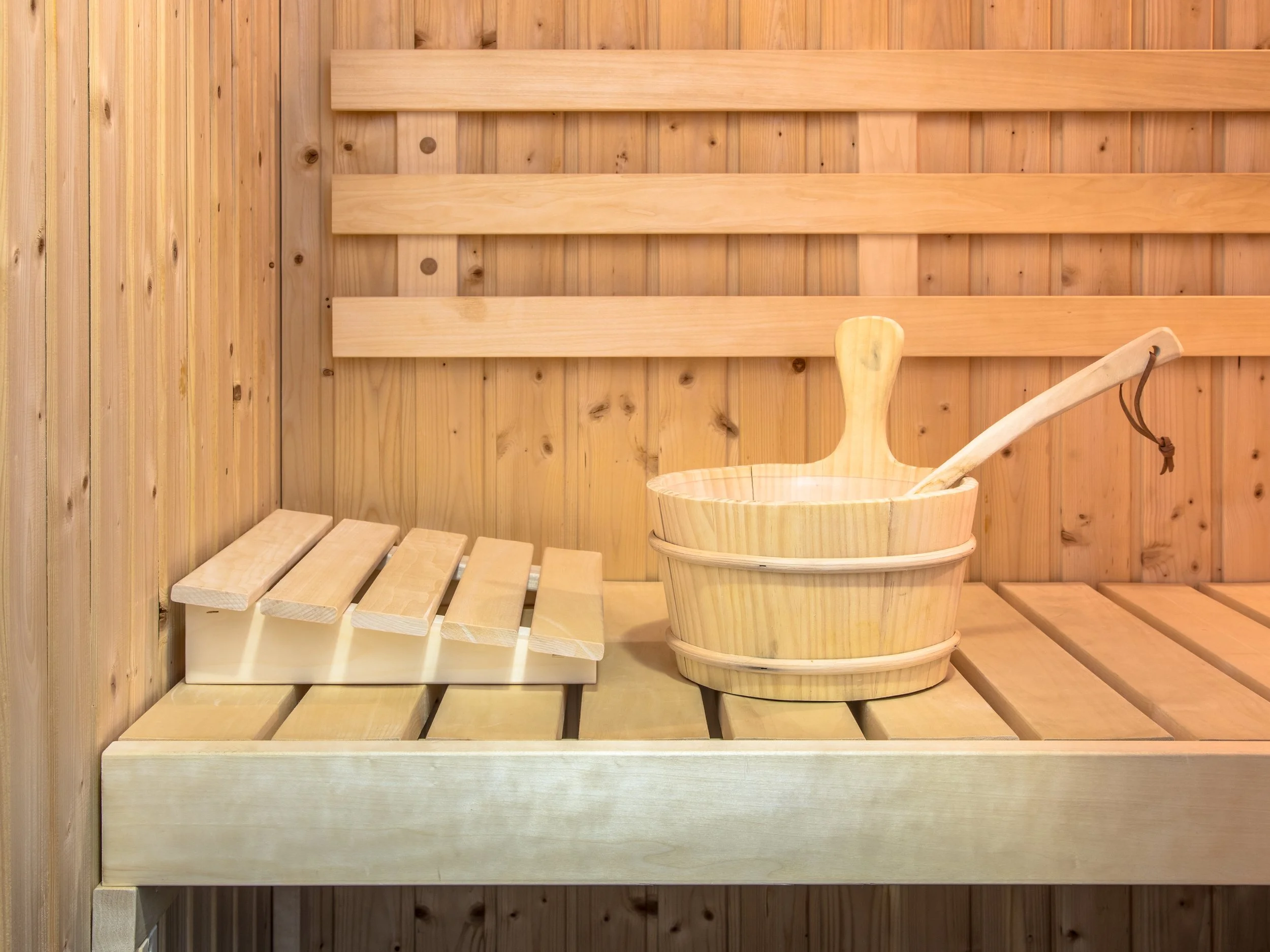 Interior of a wooden sauna with a wooden bucket and ladle used for pouring water on hot stones, and a small wooden bench.
