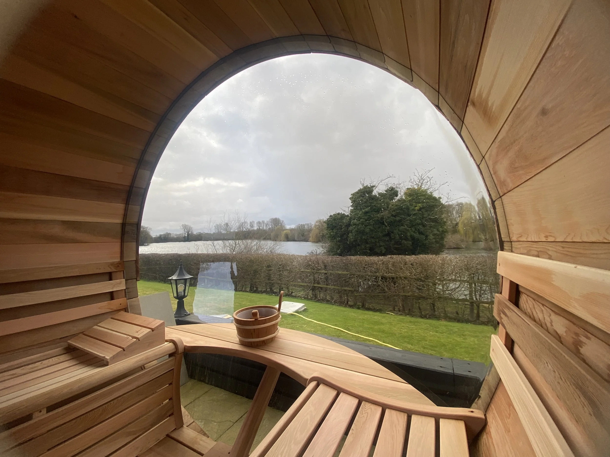 Inside a wooden barrel sauna with a large round window overlooking a lake, trees, cloudy sky, and a hedge outside.