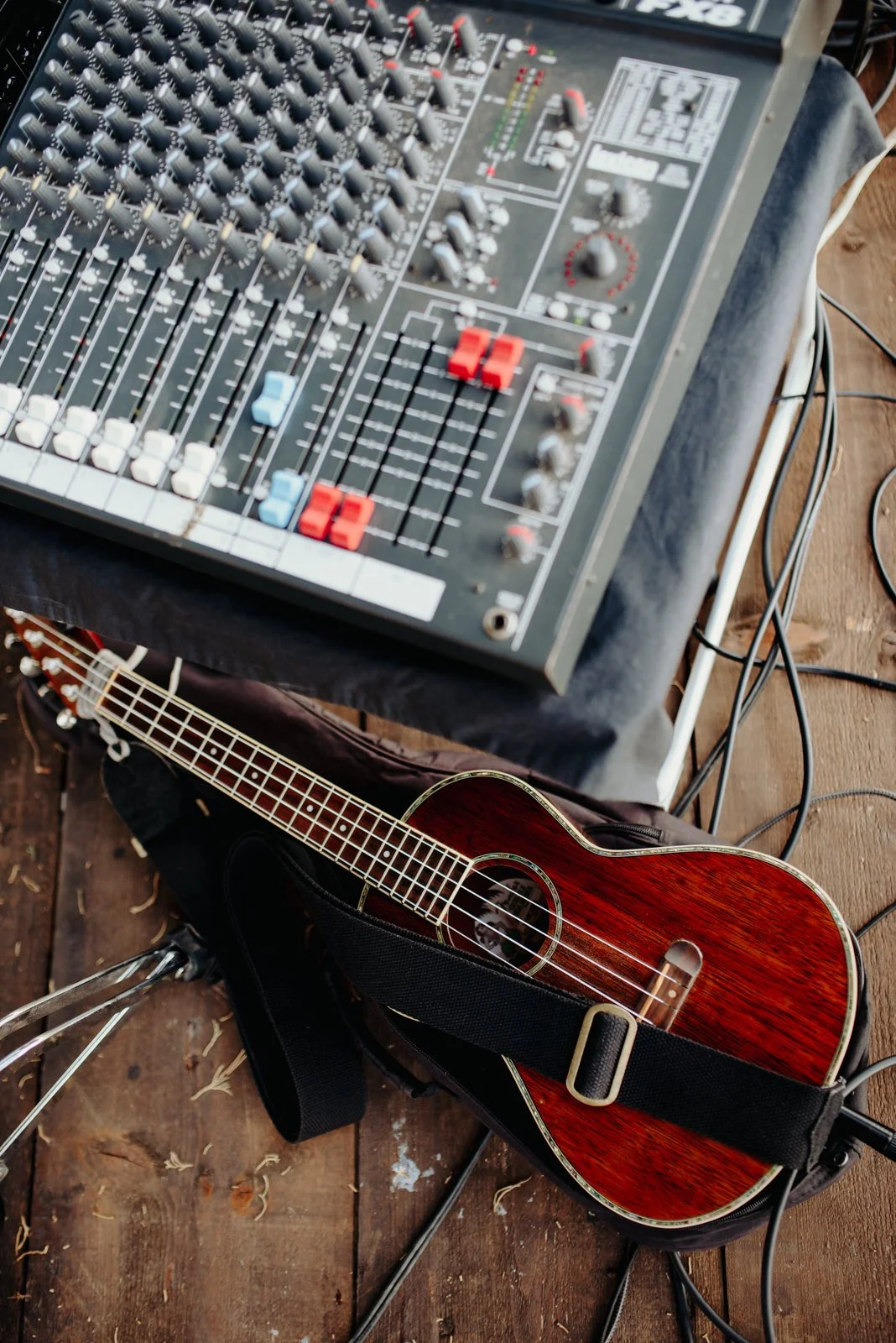 Un mixer audio con molti controlli e fader, e una chitarra acustica con una custodia, posizionati su un pavimento di legno degli Akira Manera