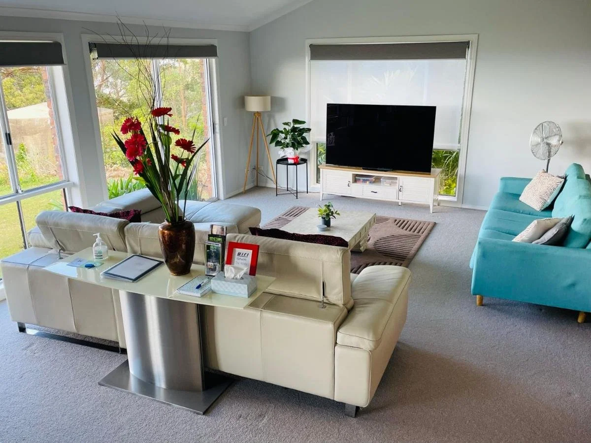 Living room with a white sectional sofa with a large floral vase, a turquoise sofa, a wooden coffee table, a TV on a white stand, and large windows with outdoor views