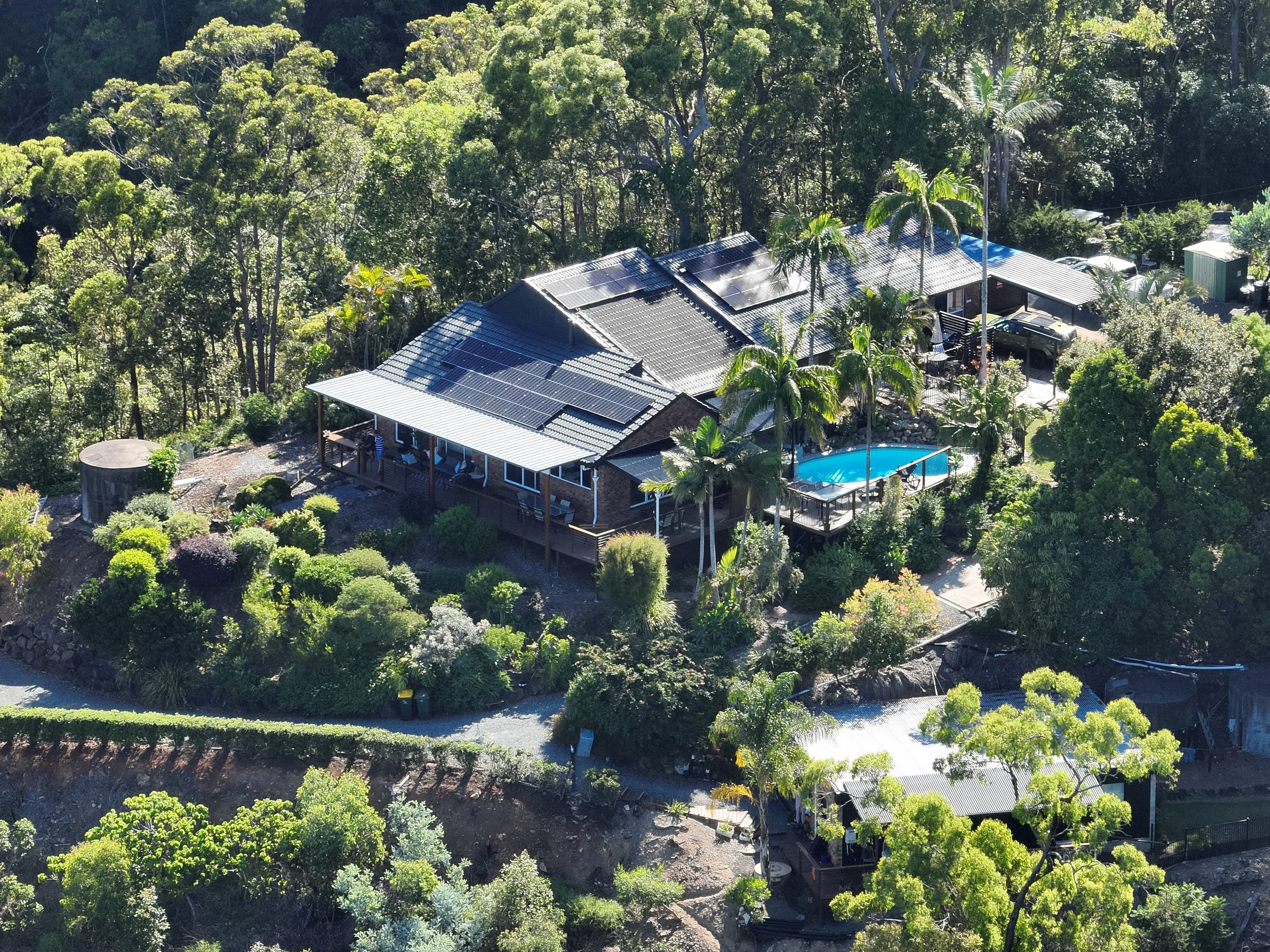 A house on a hillside surrounded by lush trees with solar panels on the roof, a swimming pool, and landscaped garden.