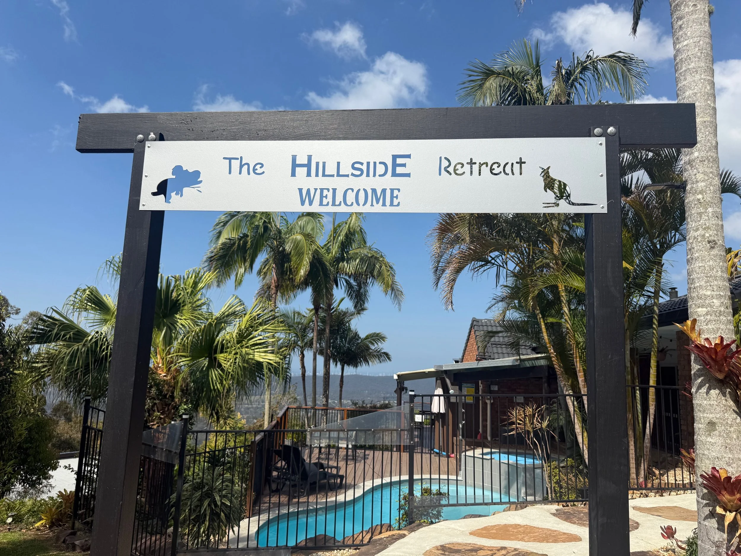 Sign that reads 'The Hillside Retreat Welcome' with a cartoon fish on the left and a cartoon kangaroo on the right, overlooking a backyard with a swimming pool, palm trees, and a clear blue sky.