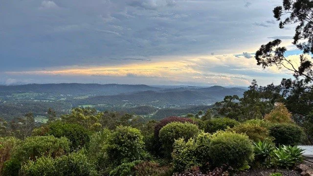 A scenic landscape with lush green bushes in the foreground and rolling hills in the distance under a cloudy sky with hints of sunlight.