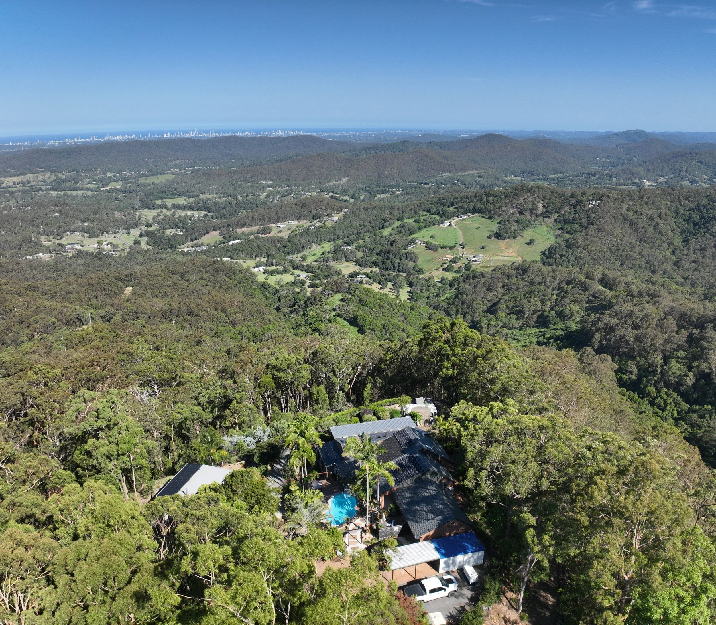Aerial view of a wooded hillside with a house, palm trees, swimming pool, and parking lot surrounded by dense trees, with rolling hills and distant city skyline in the background.