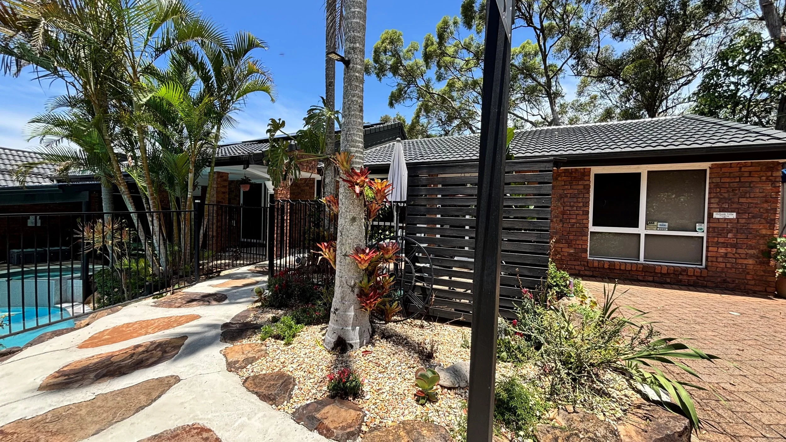 Residential backyard with a small swimming pool, tropical plants, a brick house with black-framed windows, a wooden privacy fence, and a paved patio area, under a bright blue sky.