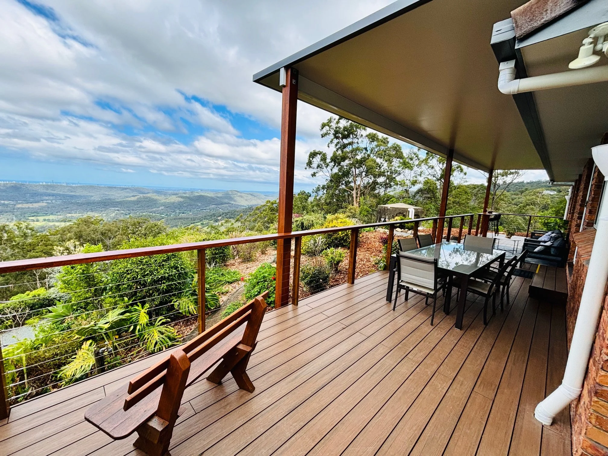 View from a wooden deck with outdoor furniture overlooking a lush green landscape with hills, trees, and a partly cloudy sky in the distance.