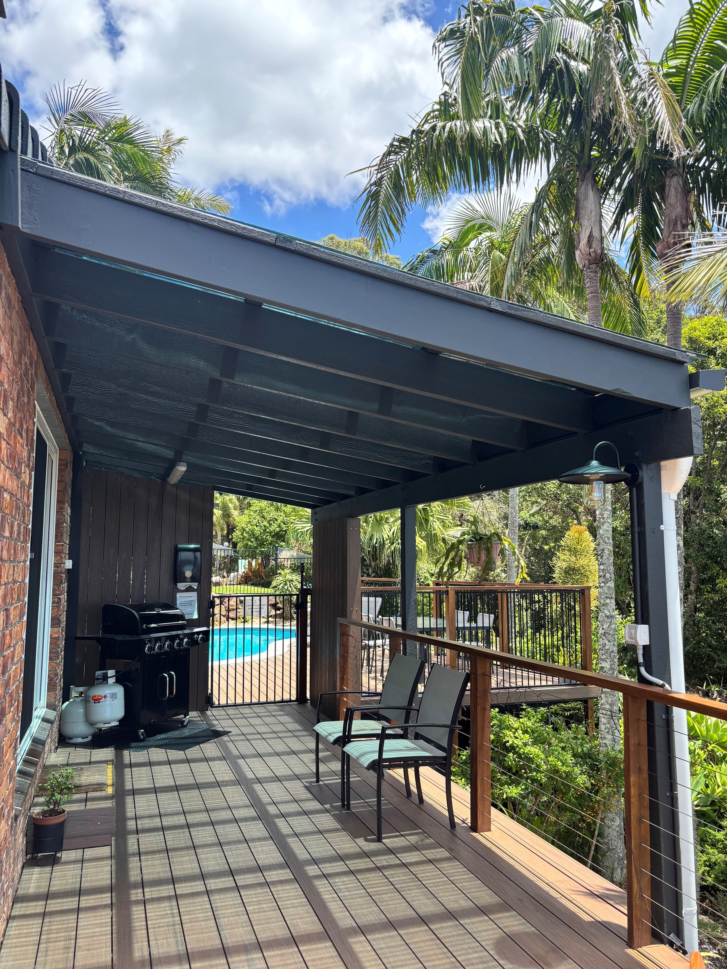Outdoor patio with chairs, a barbecue grill, a pool, lush green trees, and a partly cloudy sky.
