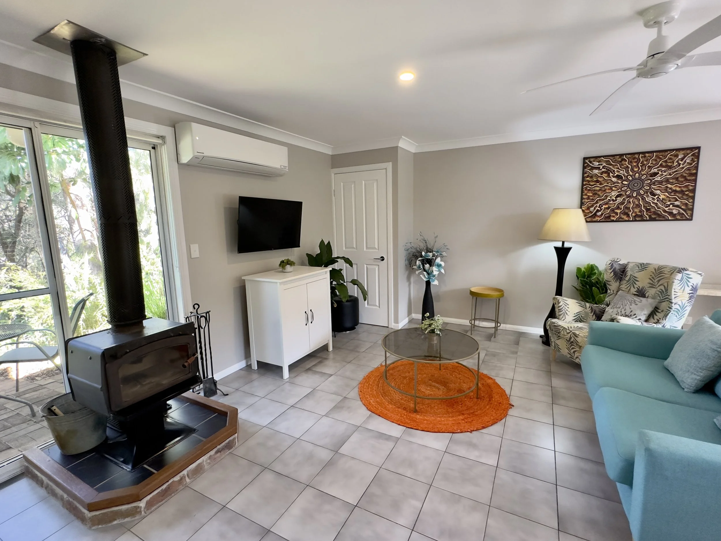 Living room with a wood stove, sliding glass door, wall-mounted TV, white cabinet, potted plant, armchair with patterned fabric, teal sofa, floor lamp, abstract artwork, coffee table on an orange rug, decorative plants, and ceiling fan.