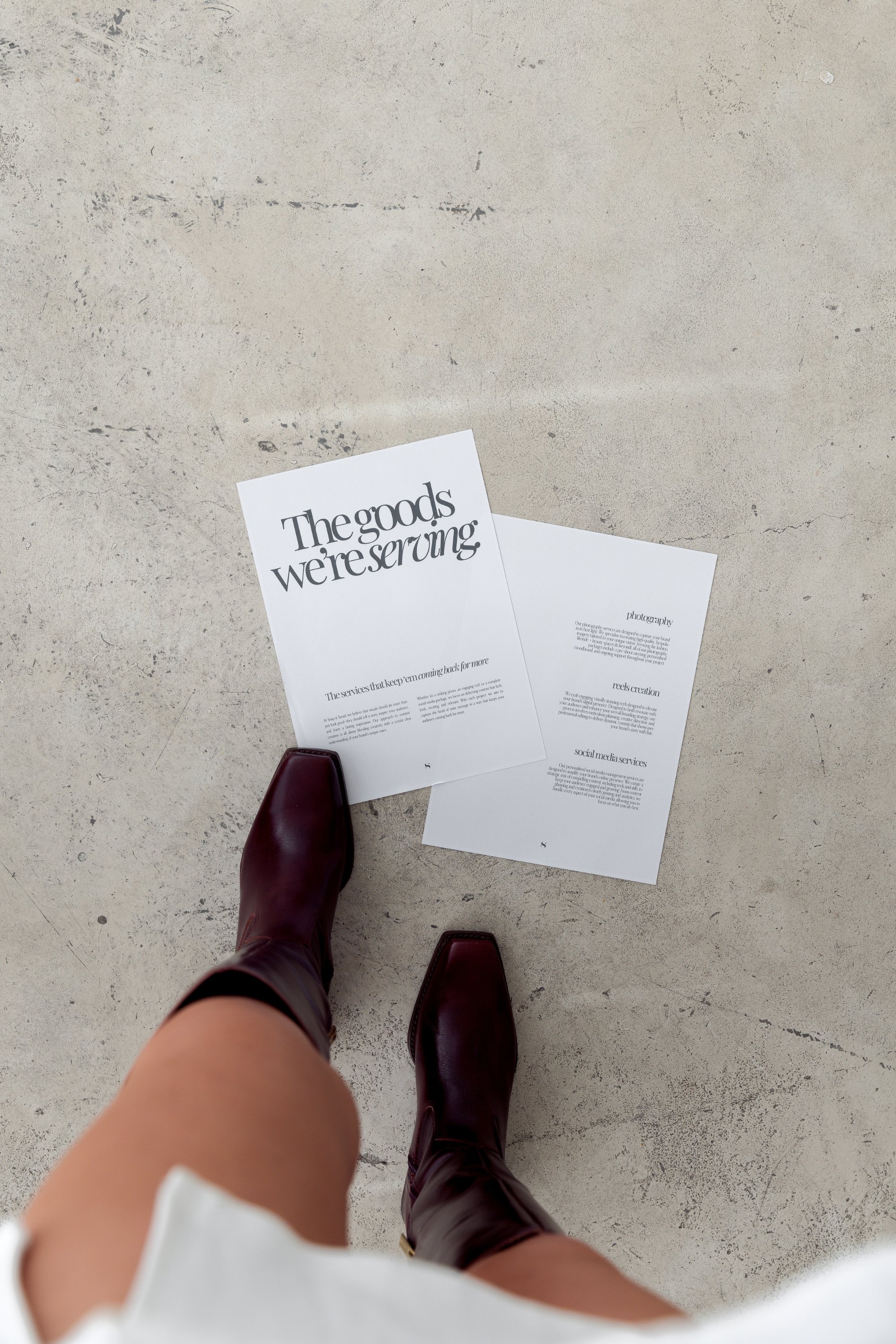 Overhead view of a person wearing burgundy boots, standing next to two printed pages on a textured floor. The visible page reads "The goods we're serving."