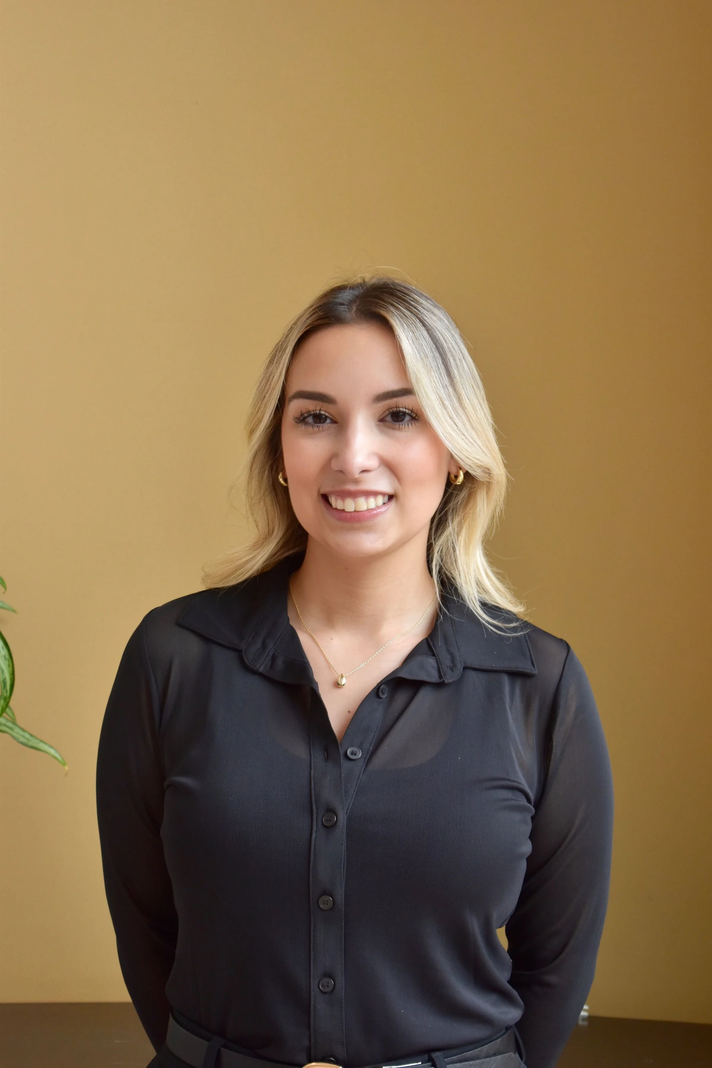 A young woman with blonde hair styled in loose waves, wearing a black collared shirt, gold hoop earrings, and a gold necklace, smiling at the camera against a plain yellow background.