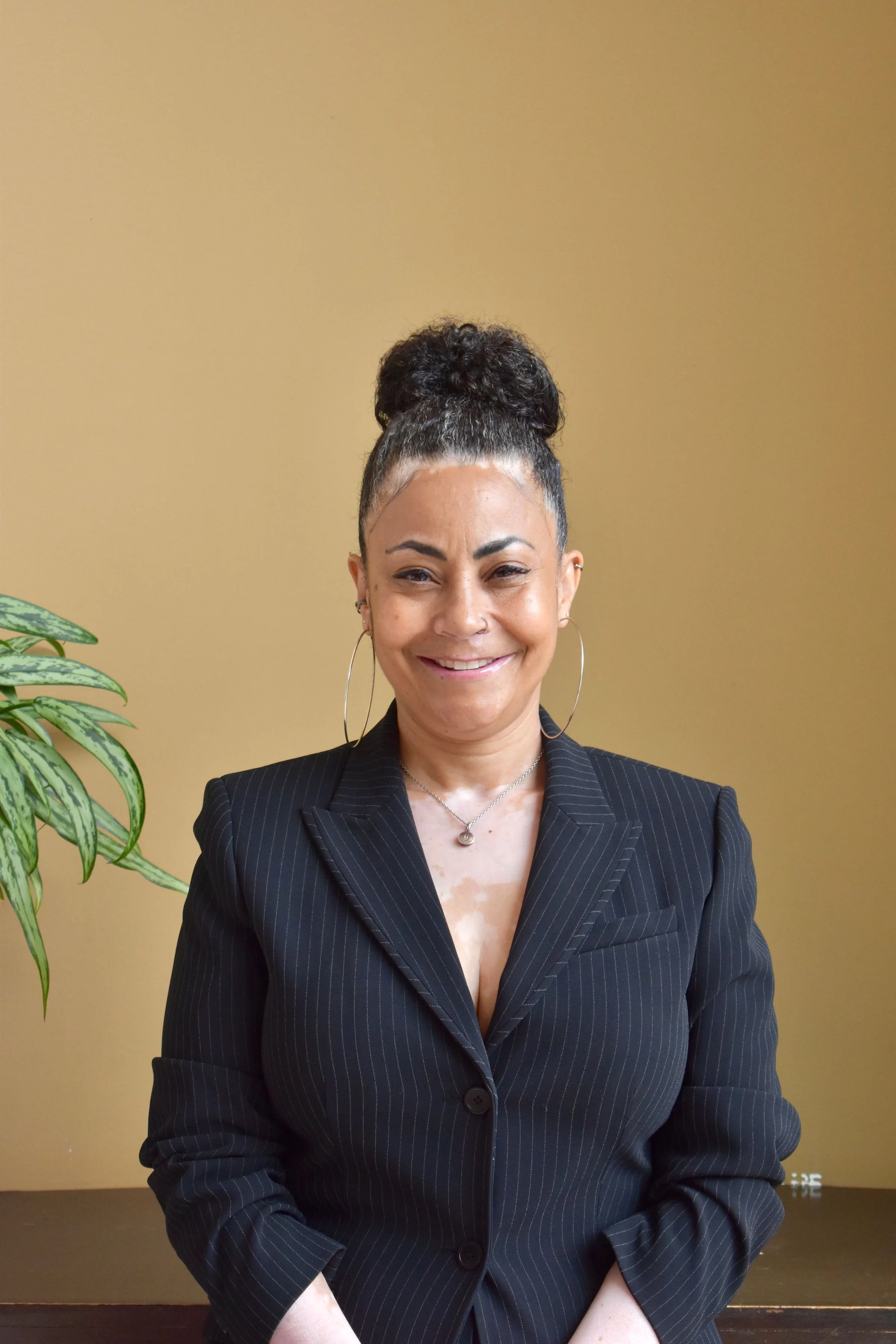 A woman with dark, curly hair tied up in a bun, smiling, wearing a black pinstripe blazer, standing against a yellow wall with a green plant on the left side.
