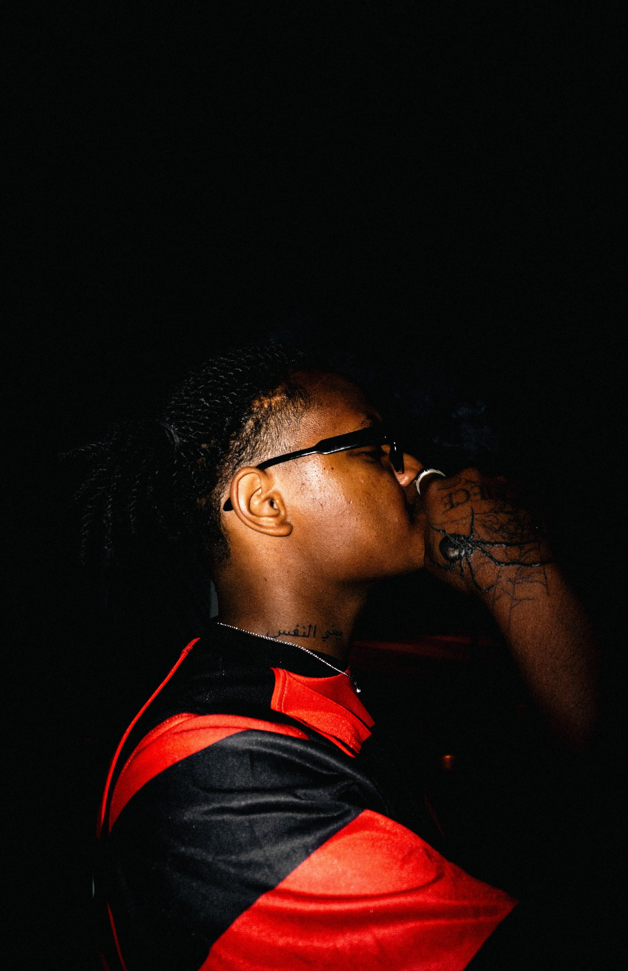 Jeune homme avec lunettes et tatouages, portant un sweat rouge et noir, en train de boire dans un verre dans l'obscurité.