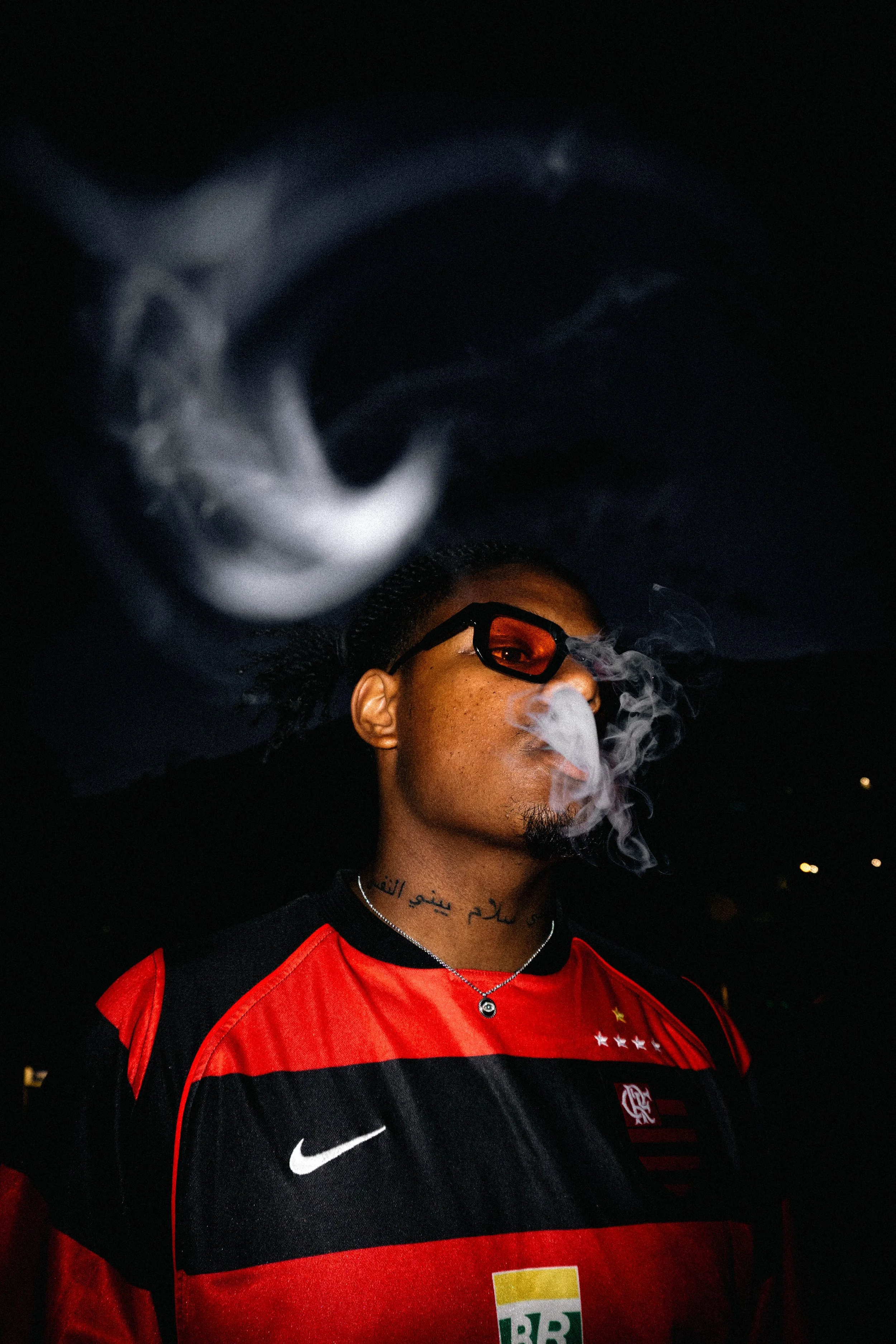 Un homme portant des lunettes de soleil, un maillot de football rouge et noir, et une chaîne avec un pendentif. Il fume de la vapeur ou de la fumée dans un environnement sombre.