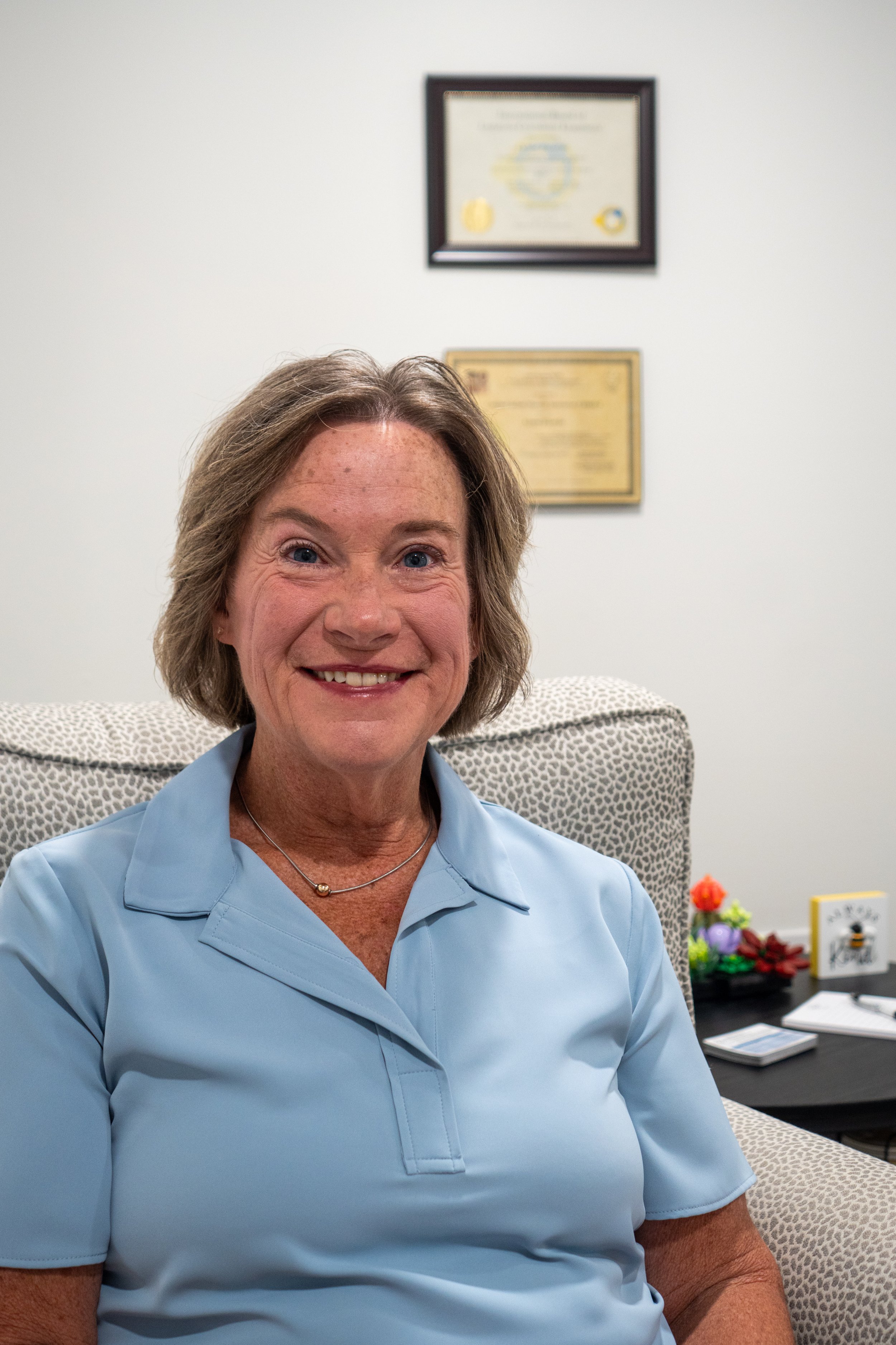 A smiling middle-aged woman with short brown hair, wearing a light blue polo shirt, sitting in an office chair in a professional office setting, with framed certificates on the wall behind her and a table with flowers and paperwork to her side.