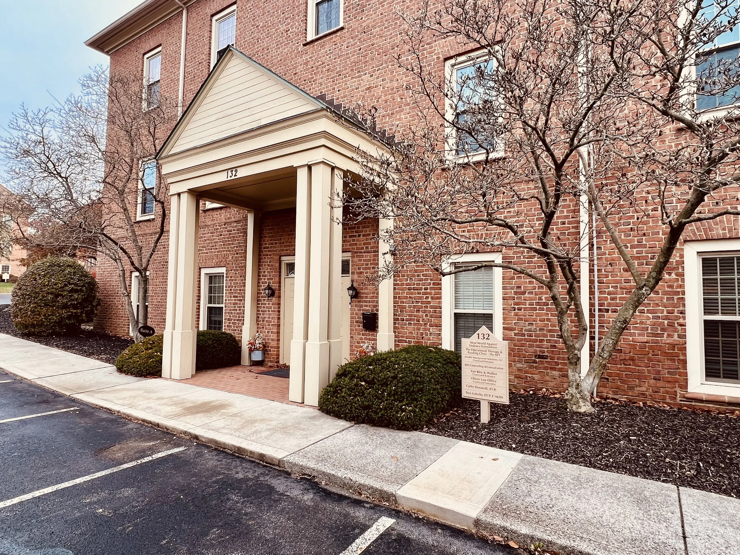Brick apartment building with a covered entrance, leafless trees, parked cars, and a sign near the sidewalk.