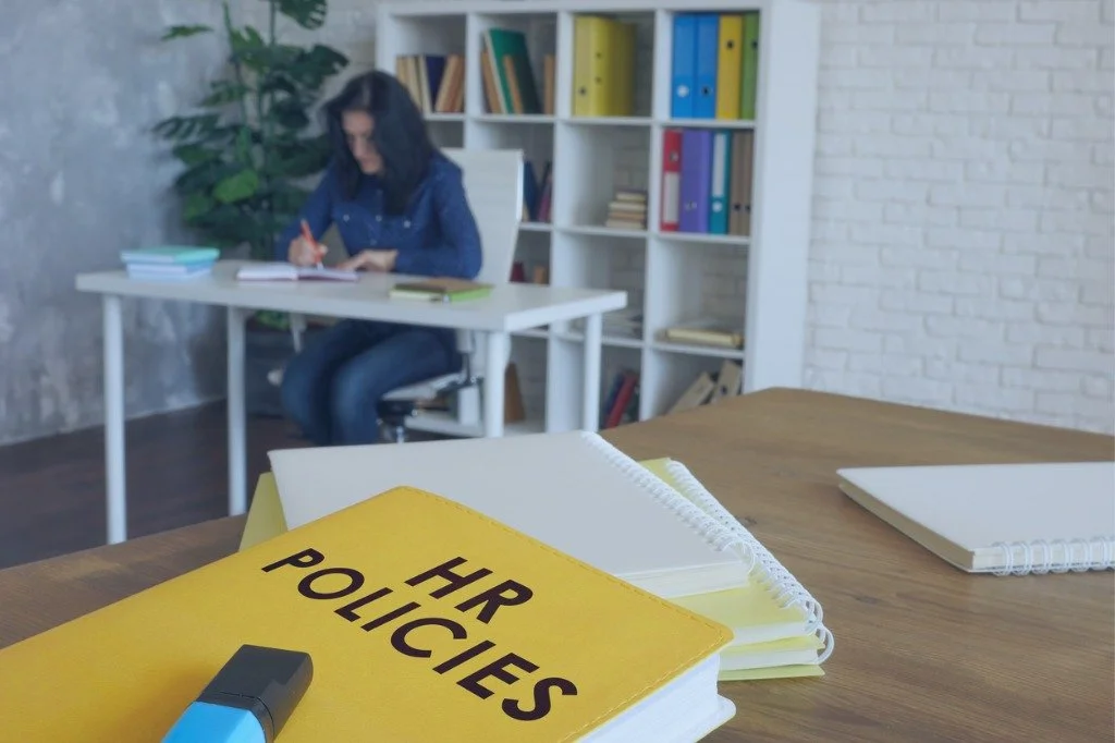 A yellow HR Policies book and some notebooks are on a wooden table. In the background, a woman is sitting at a desk writing, with a bookshelf filled with colorful folders and books behind her.