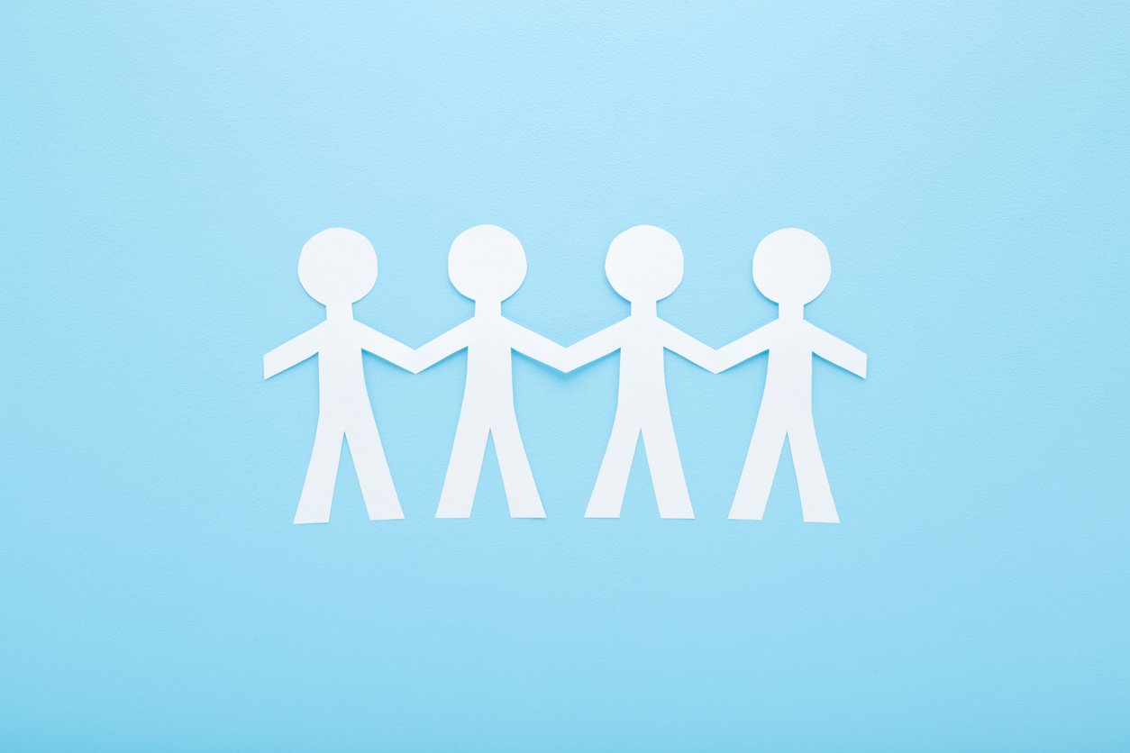 Cut human shapes from white paper on light blue table background. Pastel color. Holding hands together. Social relationship concept. Closeup. Top down view.