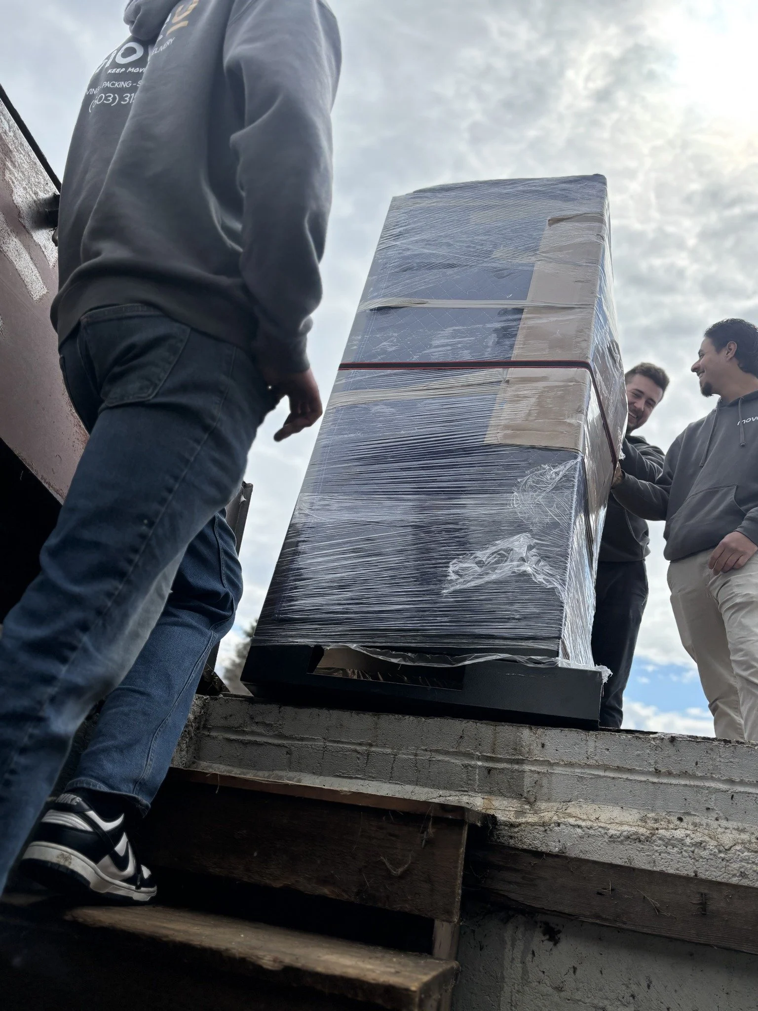 Three people loading a large wrapped box onto a truck, seen from a low angle with partly cloudy sky in the background.