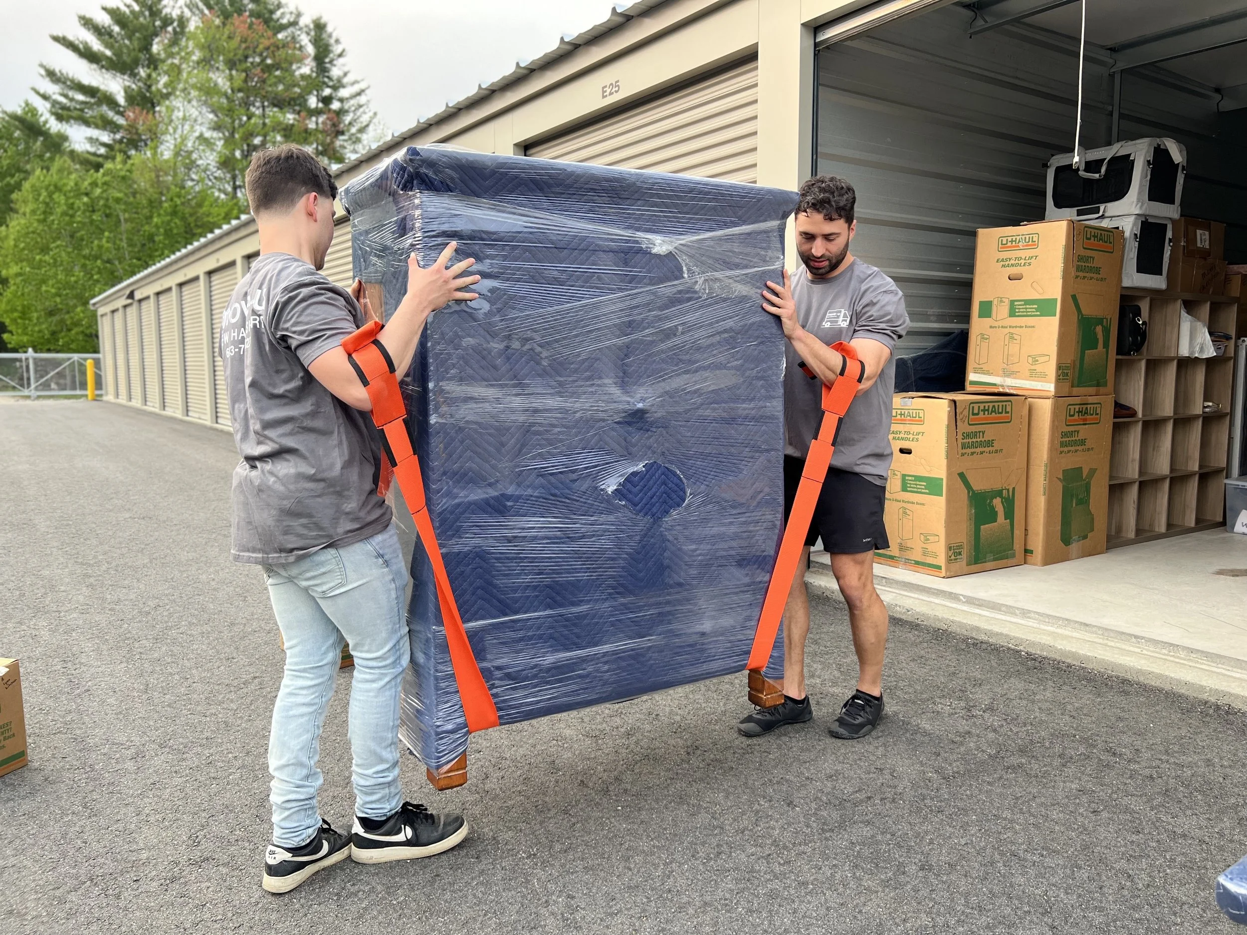 Two men in gray shirts and black shorts lifting a large boxed item wrapped in plastic outside a storage unit, with U-Haul boxes and shelves nearby.