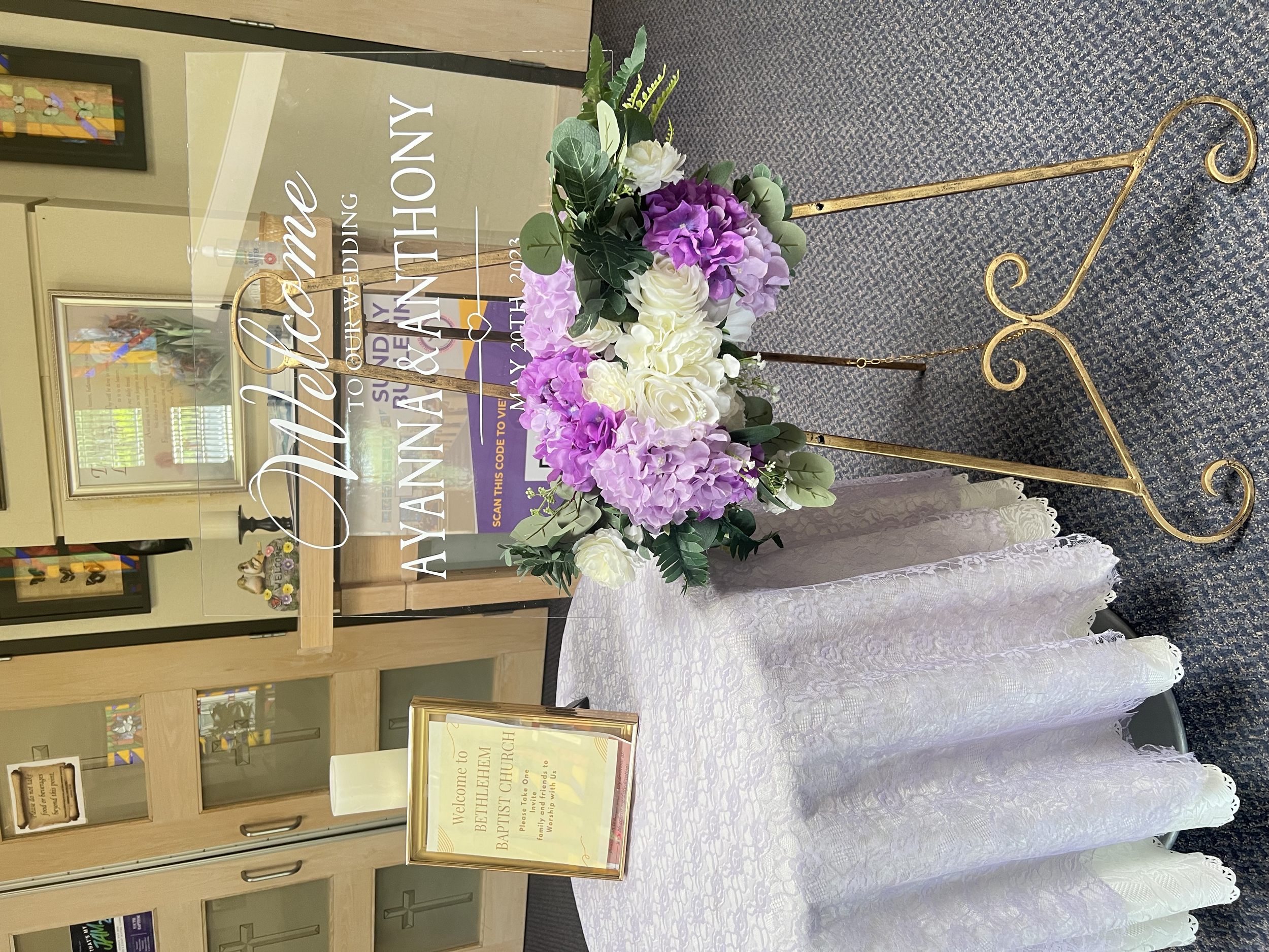 A floral arrangement with pink, white, and purple flowers on a gold stand, a white lace cloth, and framed signs welcoming guests to a wedding at Bethel Church.