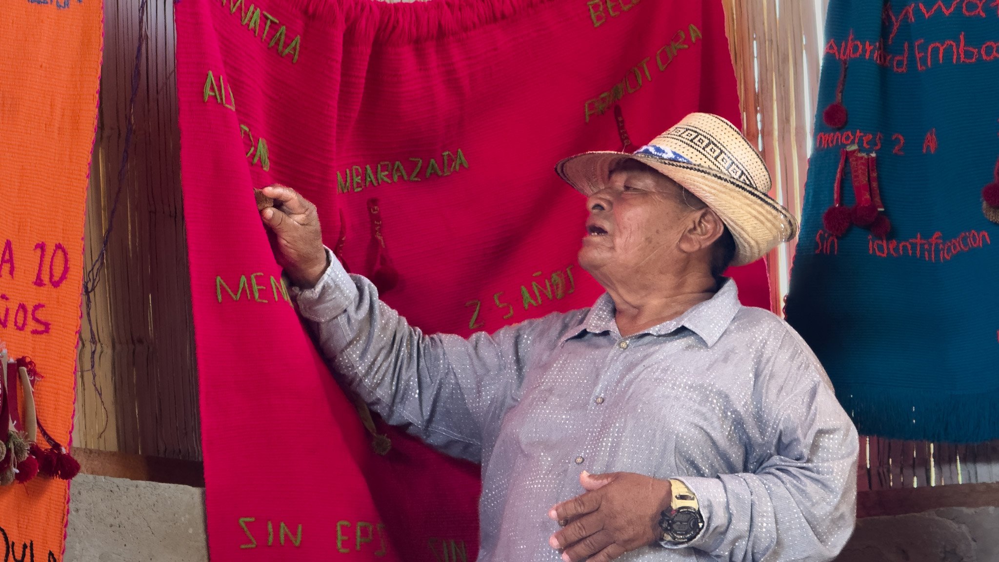 Un hombre de edad avanzada con sombrero de paja interactuando con tela de colores en un entorno cultural latinoamericano.